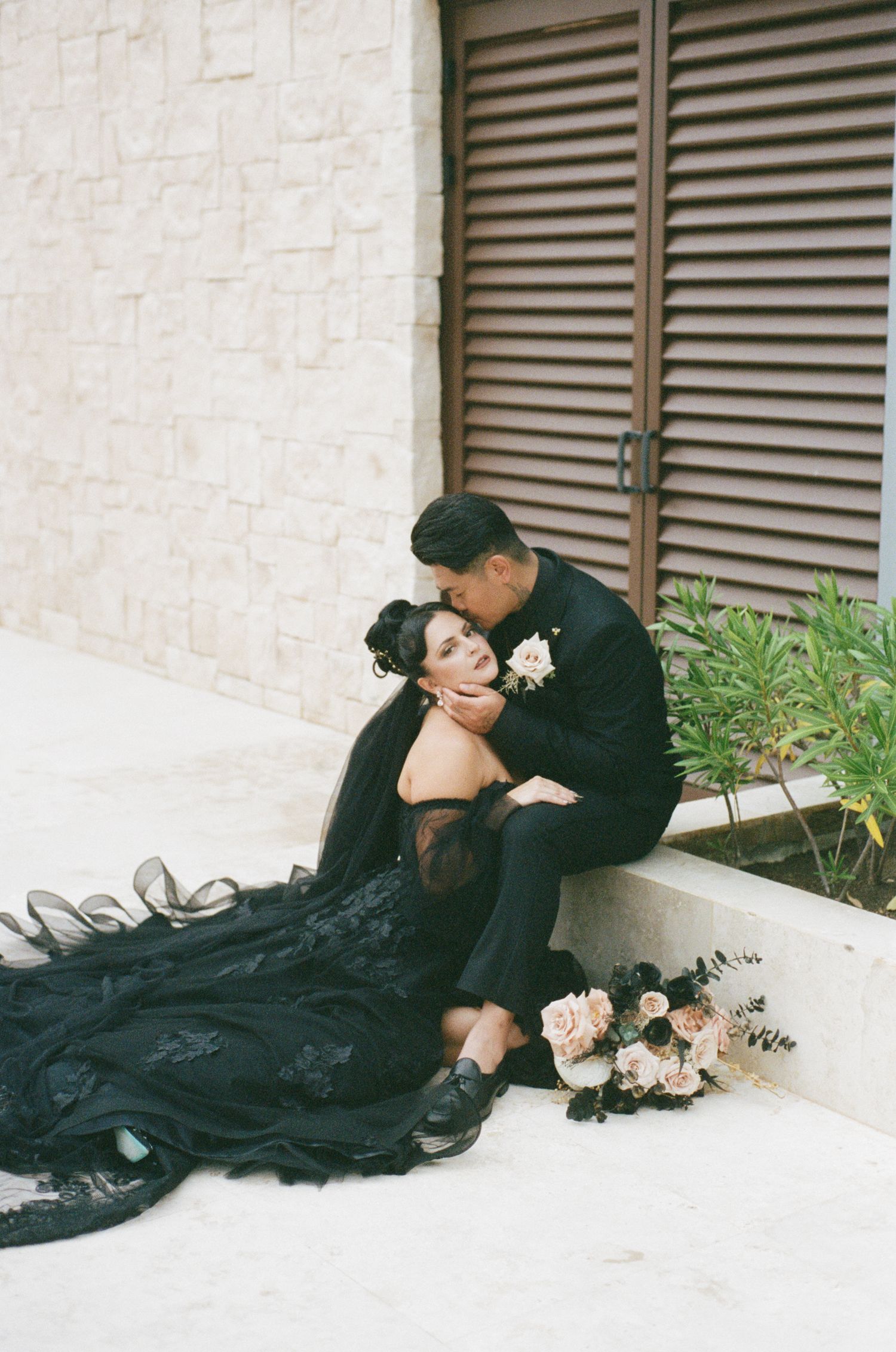 Groom in black suit kisses bride wearing black lace wedding dress while sitting on steps outdoors.