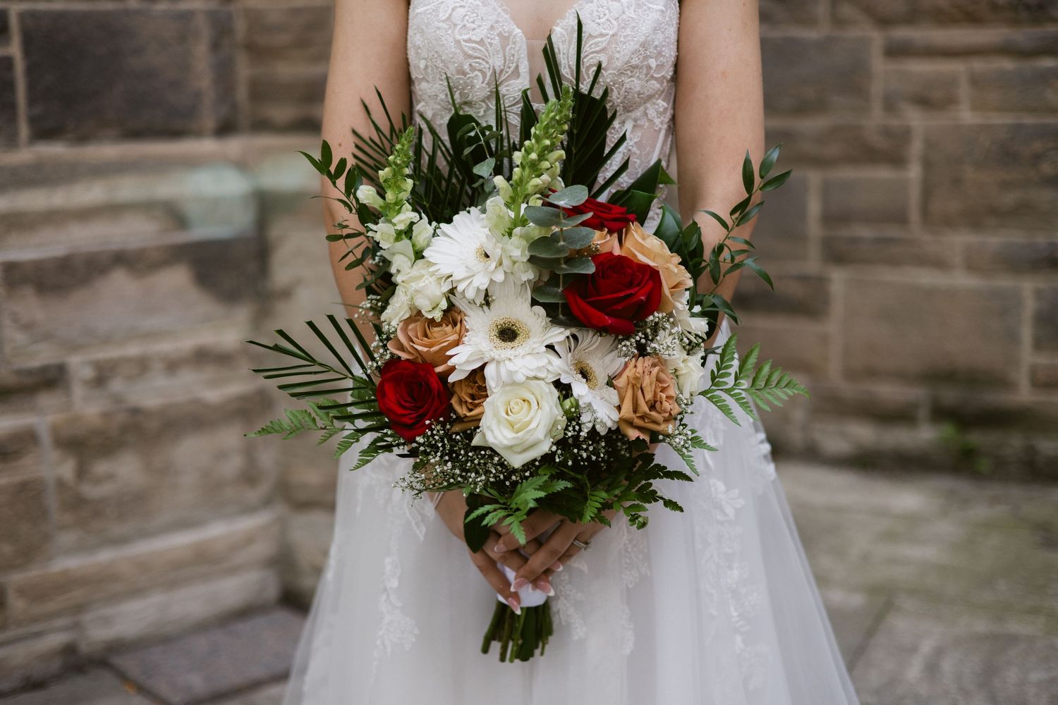 Close-up of bride holding lush wedding bouquet of red, white, and peach roses with tropical greenery.