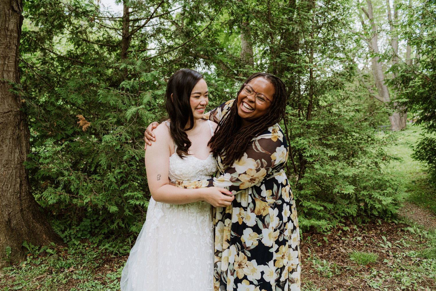 Bride in lace gown hugging and laughing with friend in floral dress at outdoor wedding.