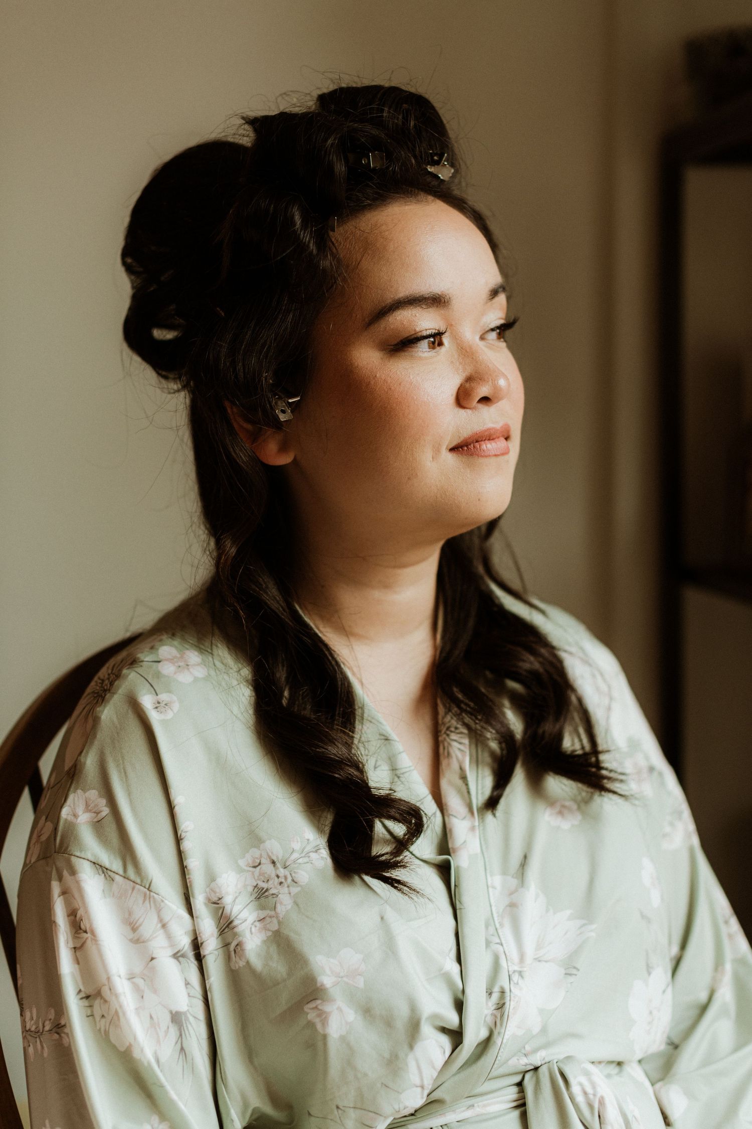Bride in floral green robe with hair in rollers gazing away during bridal prep.
