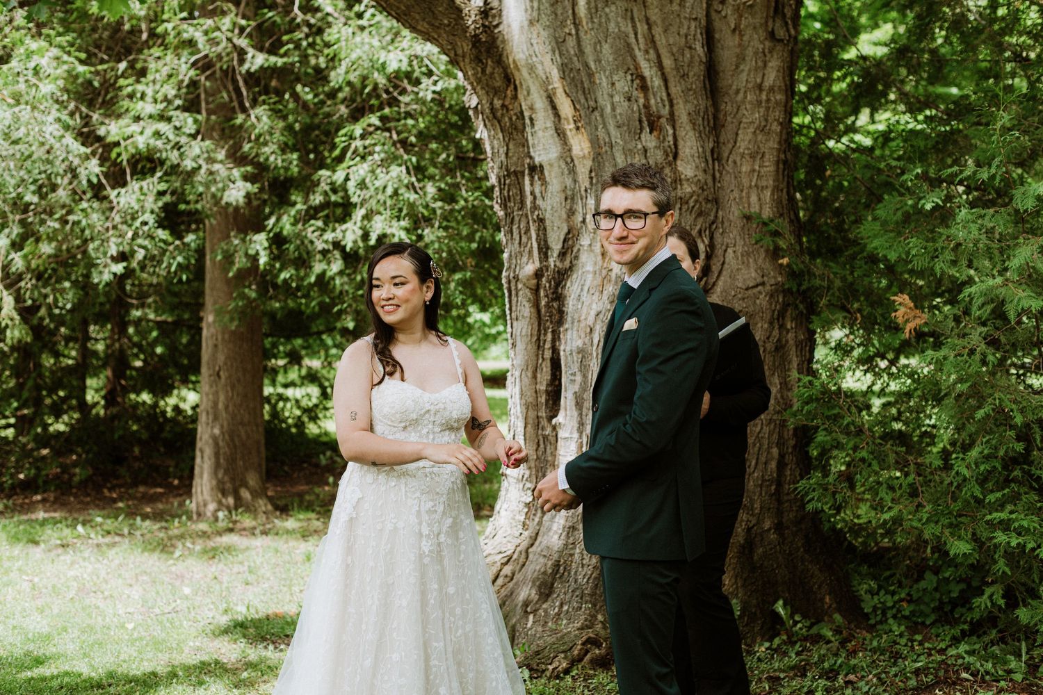 Bride and groom smile together standing near large tree after outdoor wedding ceremony in garden.