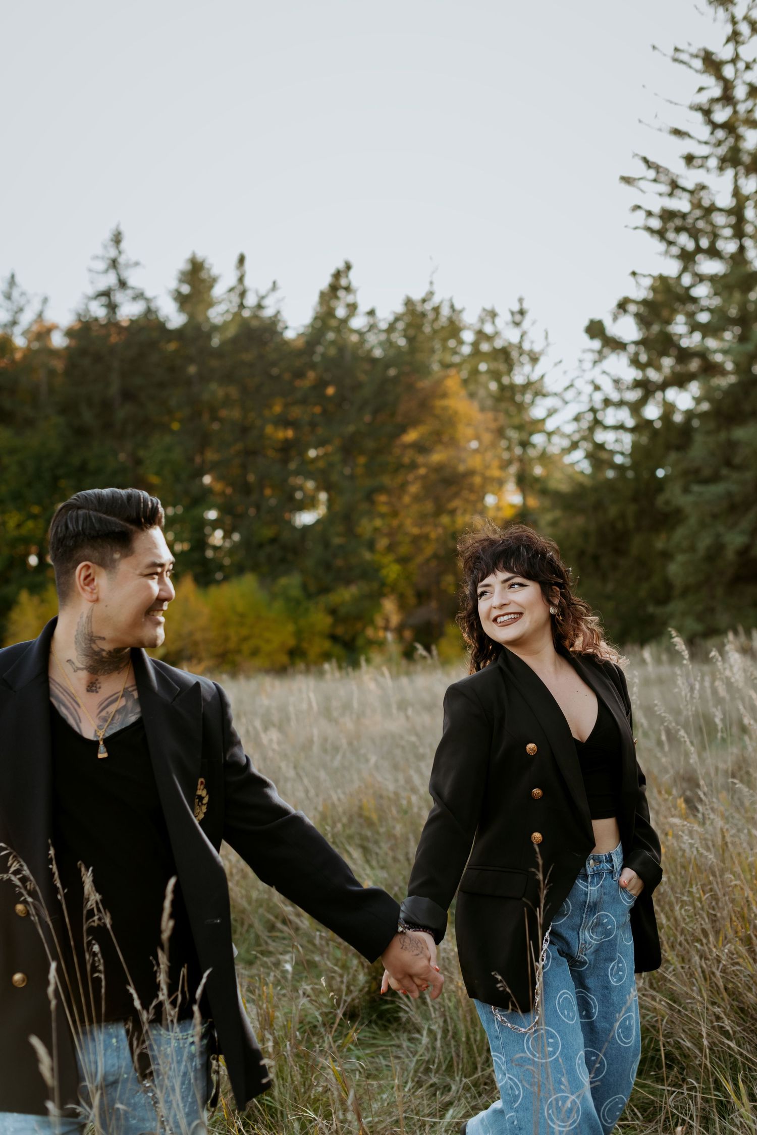 Smiling couple holding hands while walking through tall grass meadow with colorful autumn evergreen trees in background.