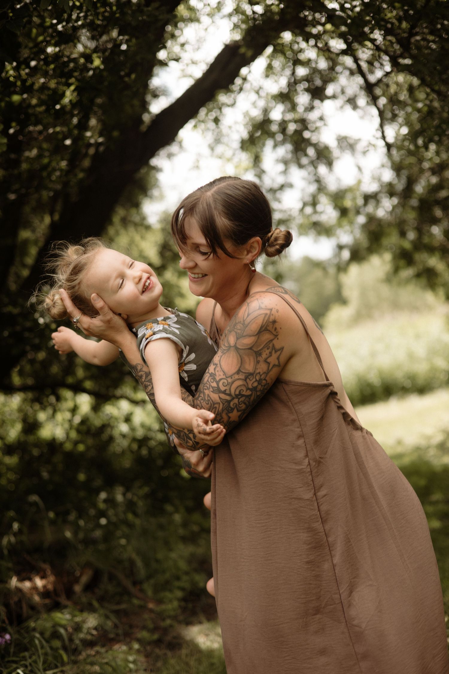 A smiling tattooed mother holds her laughing toddler in a lush green setting.