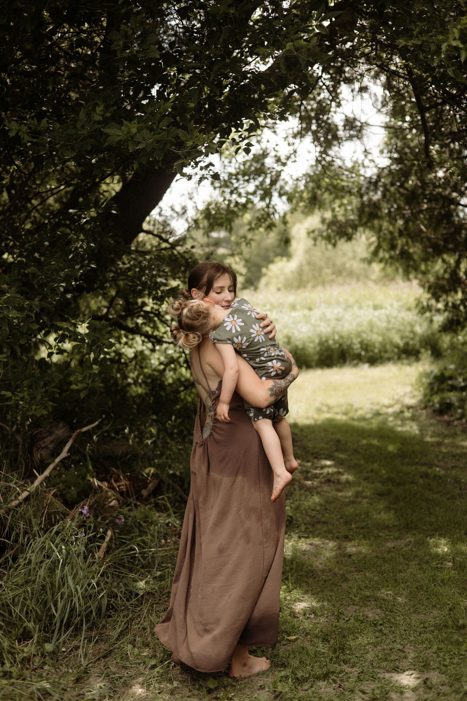 A tattooed mother in a brown maxi dress hugs her toddler child under lush green trees outdoors.