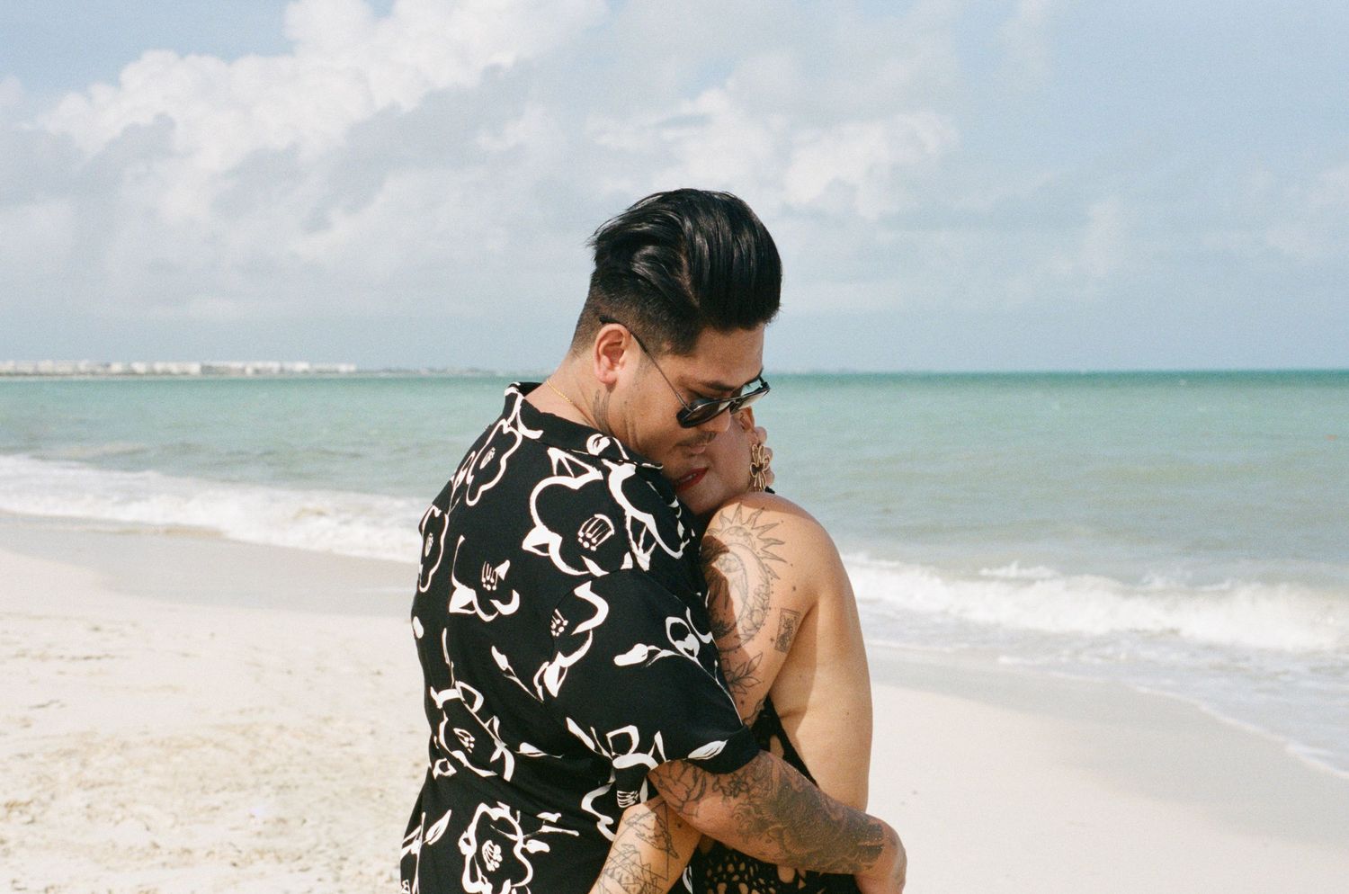Couple sharing an intimate embrace on a sunny beach, man in floral shirt kissing woman's forehead.