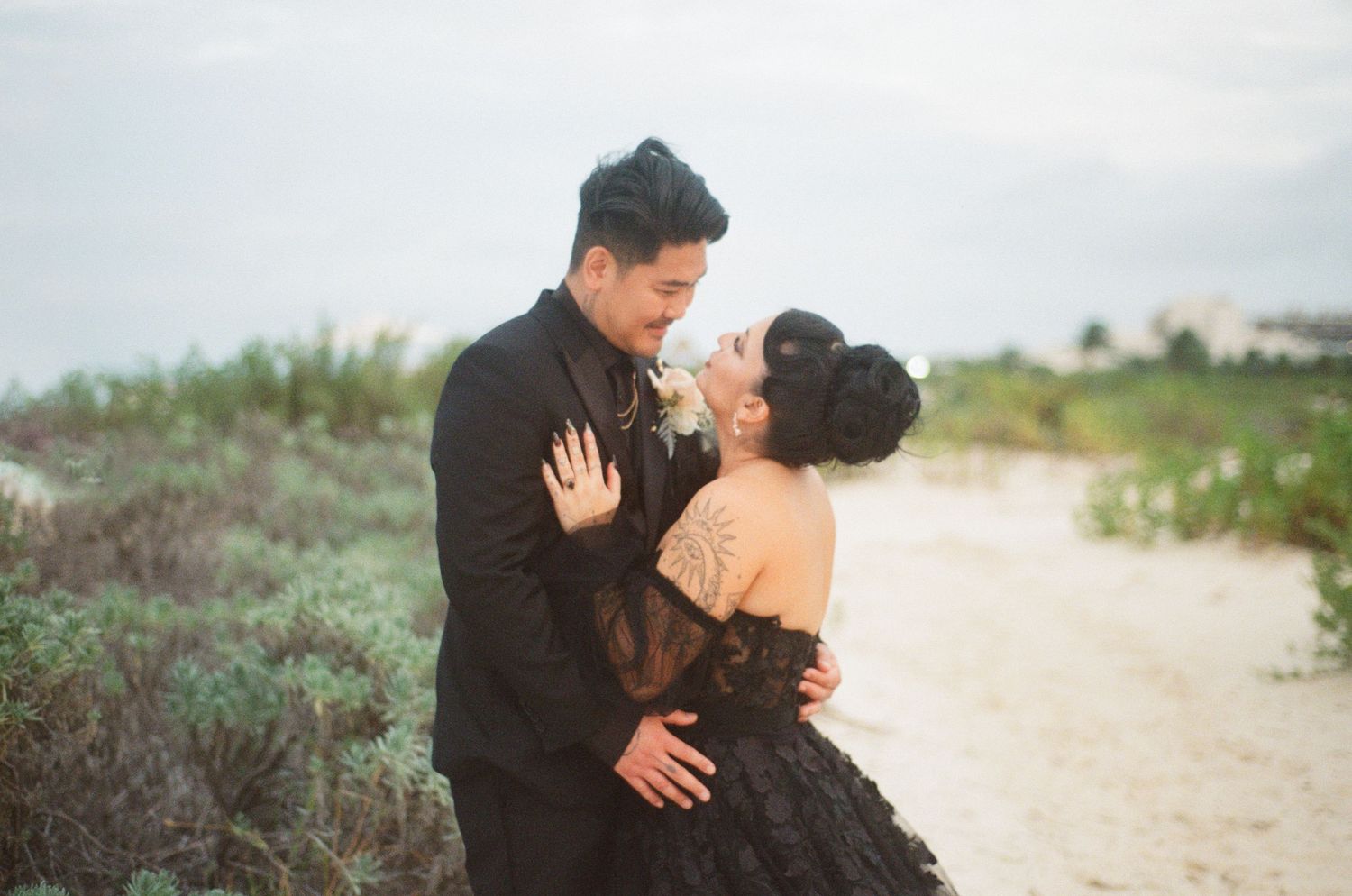 Smiling couple embracing on sandy beach path, bride in black lace gown, groom in black suit with boutonniere.
