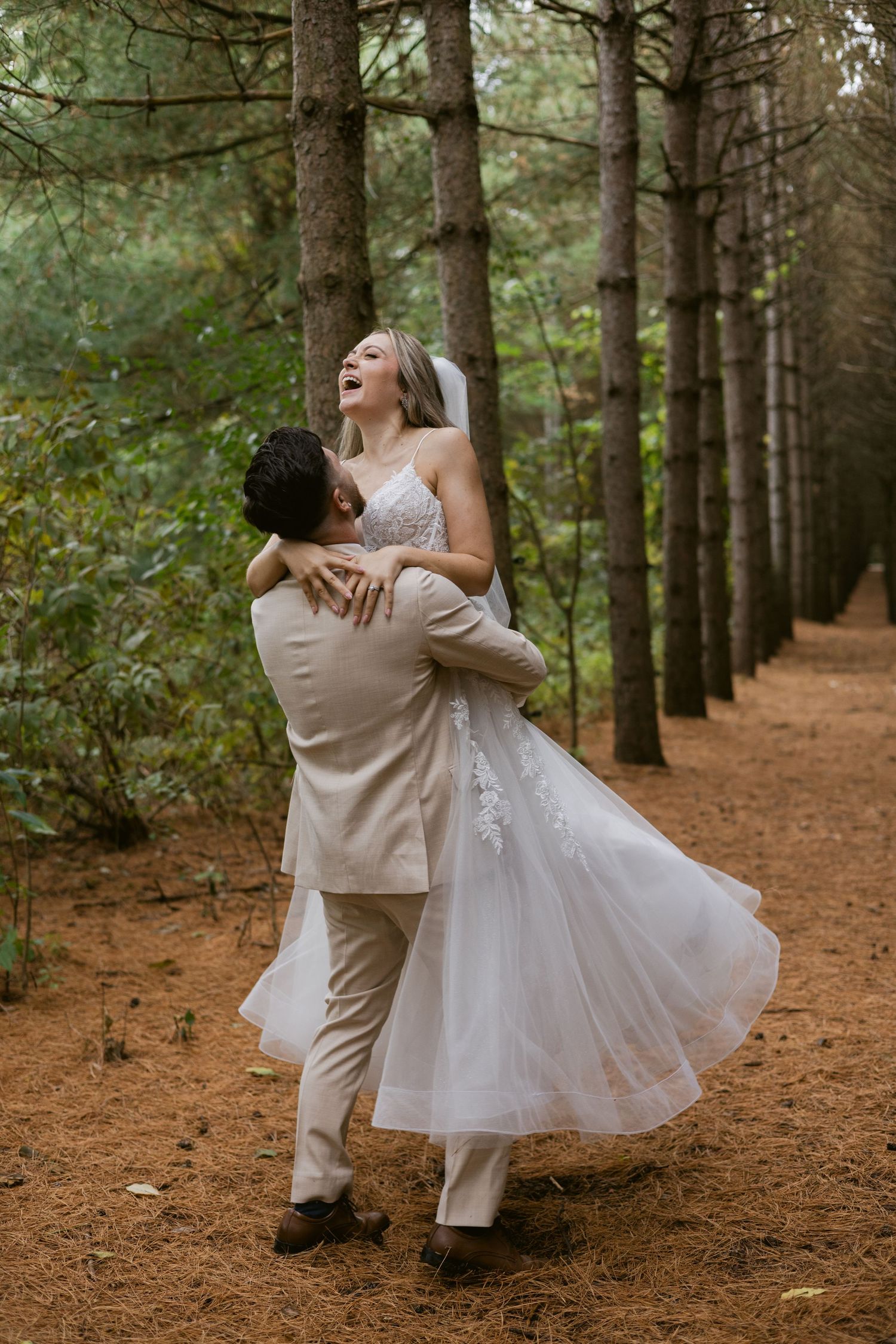 Groom lifts laughing bride in a flowing white gown along a pine forest path on their wedding day.