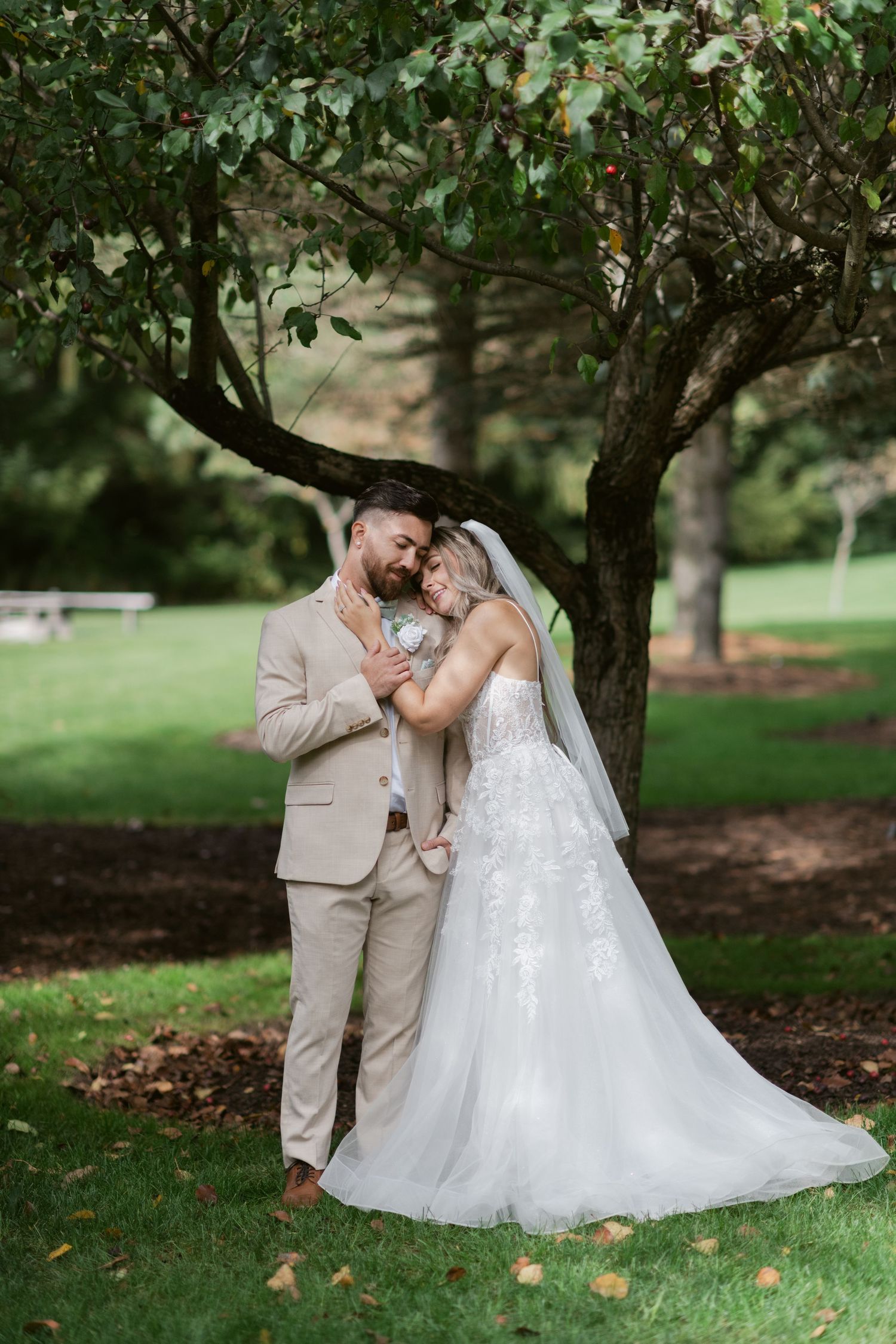 Bride and groom in tan and white wedding attire snuggling under a leafy tree in a park.