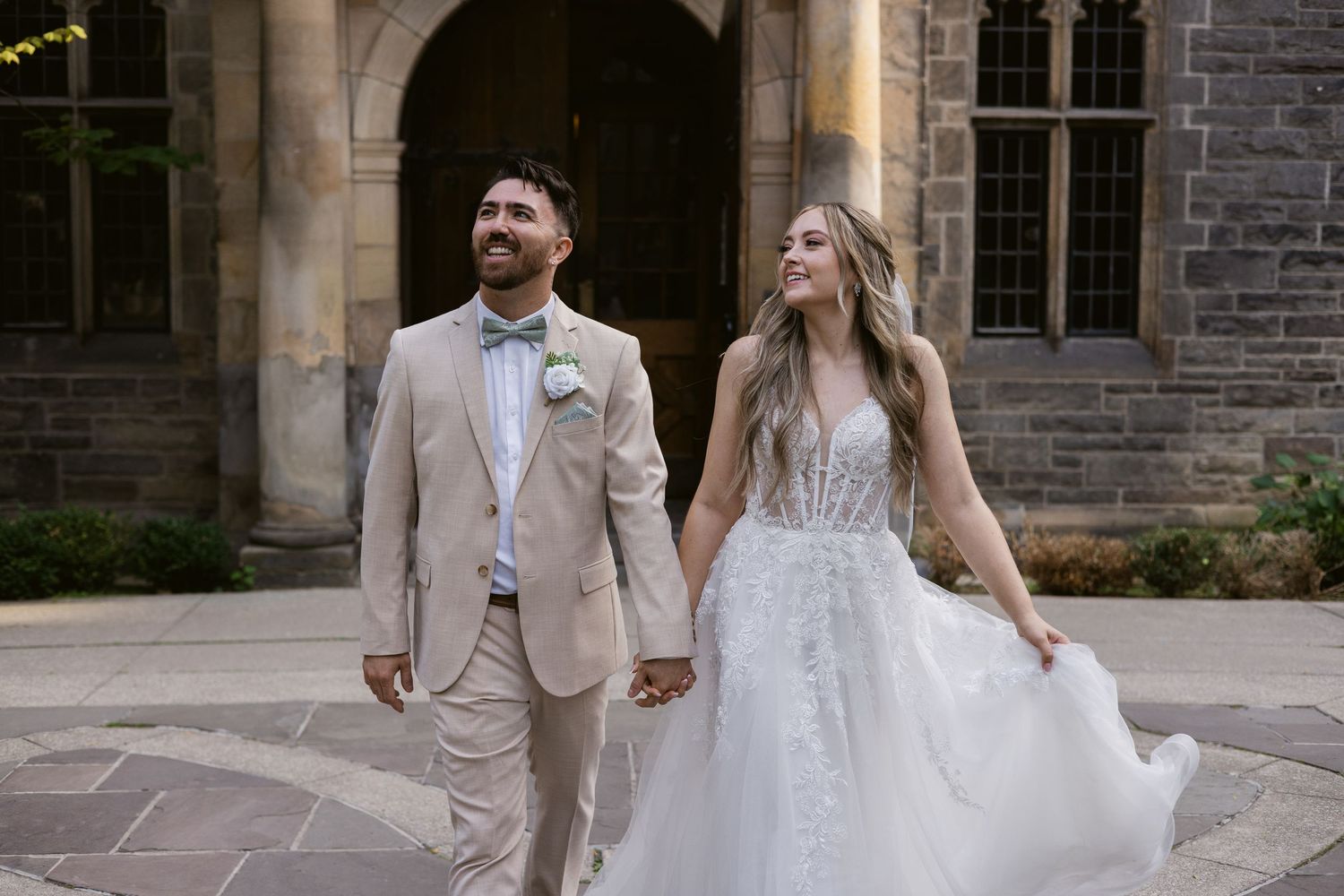 Bride and groom walk hand-in-hand smiling in courtyard of elegant stone wedding venue.