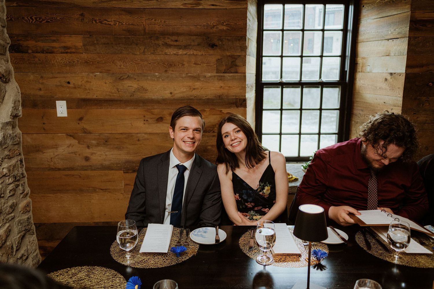 Smiling couple posing together at a wedding reception dinner table near a rustic wood-paneled window.