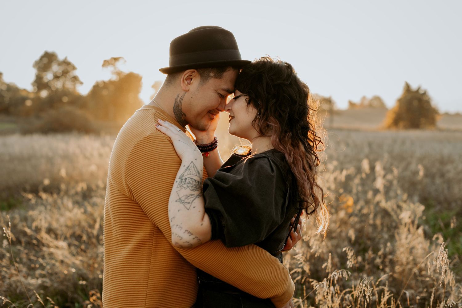 Romantic couple nearly kissing in sun-drenched field, man in mustard sweater and black hat embracing woman at golden hour.