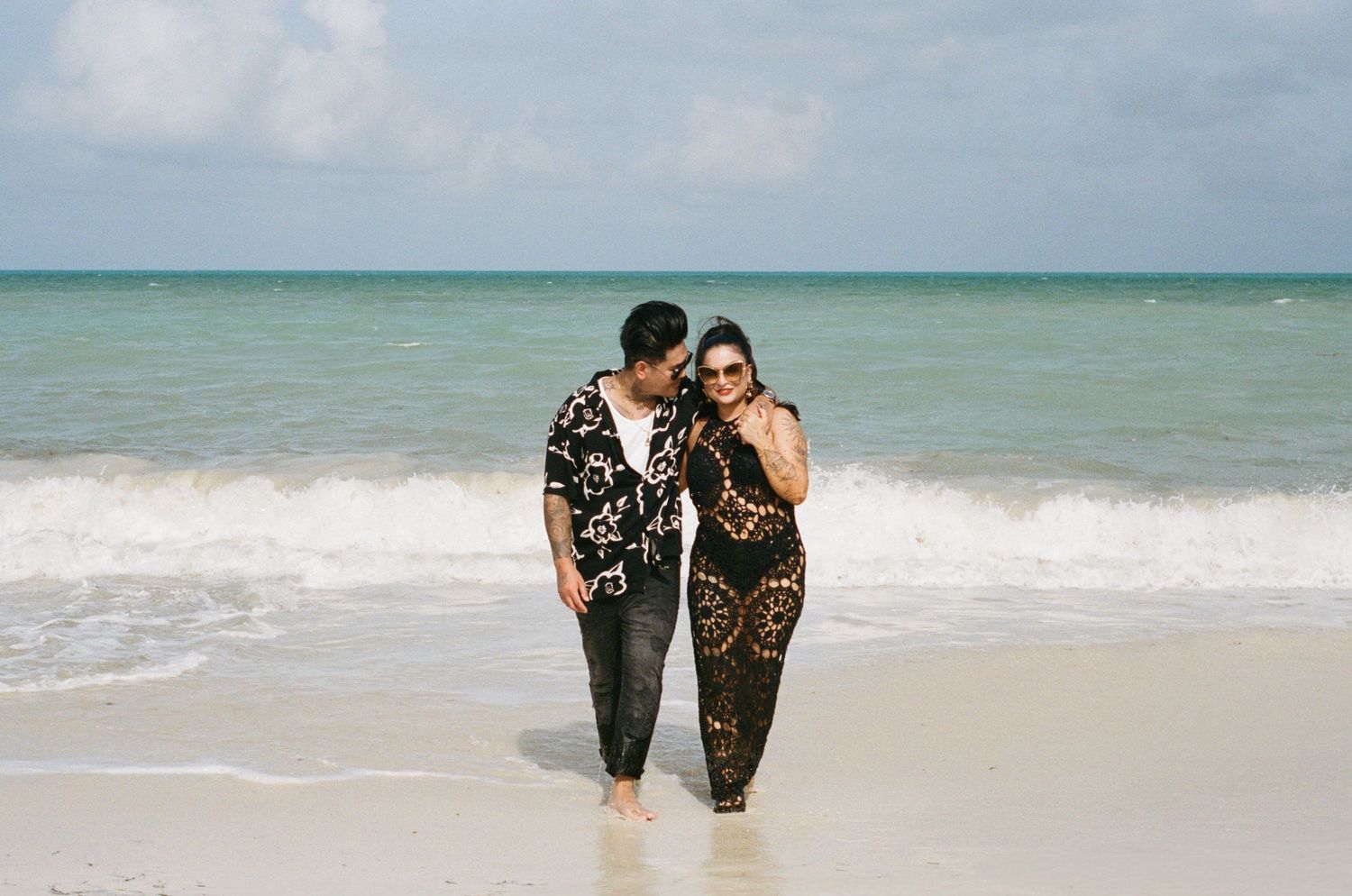 Couple in black outfits standing together at shoreline on beautiful turquoise Caribbean beach.