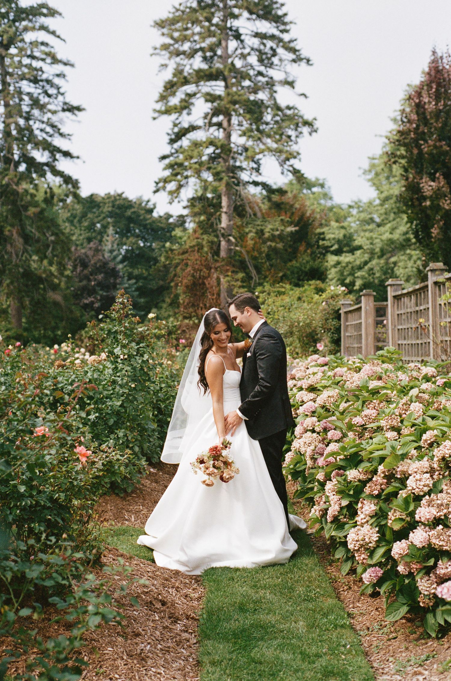 Bride and groom walking through a lush garden path surrounded by hydrangeas and roses.