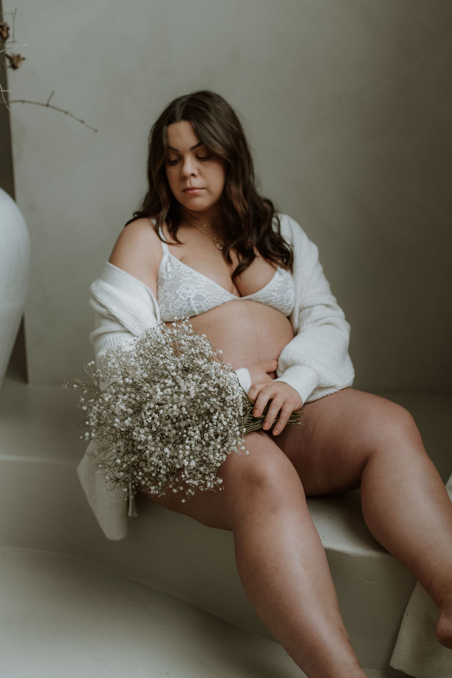 Pregnant woman in white lace lingerie holds baby's breath flowers while seated in a neutral studio.