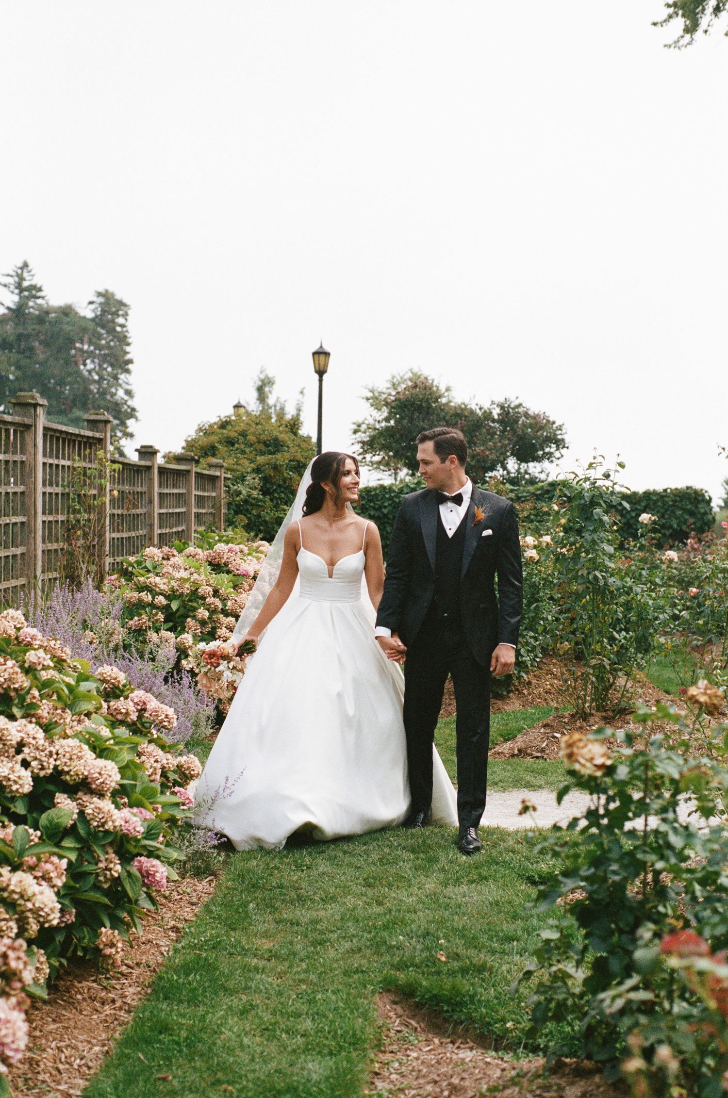 Elegant bride and groom walking hand in hand through a beautiful garden with hydrangeas.