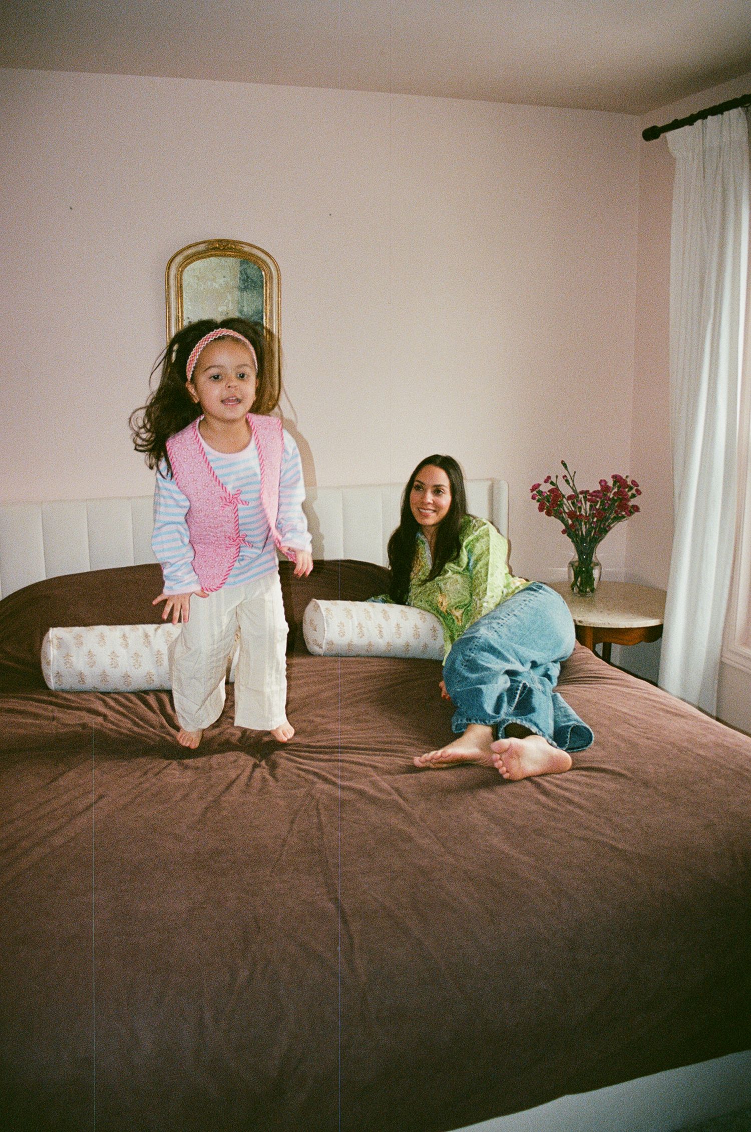 A young girl jumps on a brown bed while a woman sits smiling in a bright bedroom with pink walls.