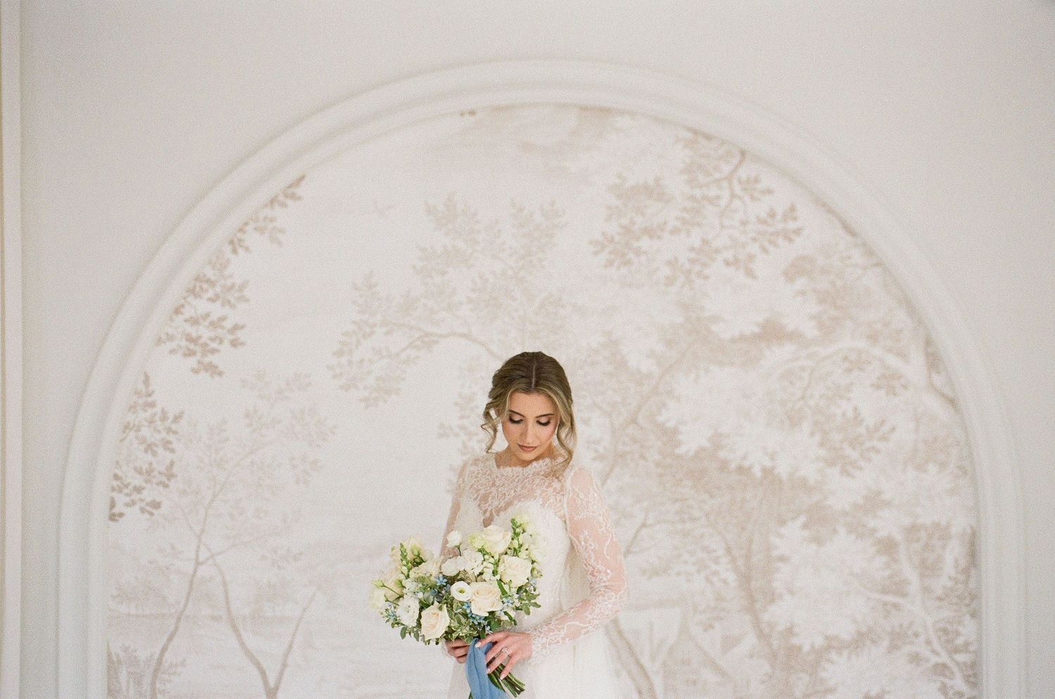An elegant bride in a lace gown holds a white bouquet standing before an ornate arched floral mural.