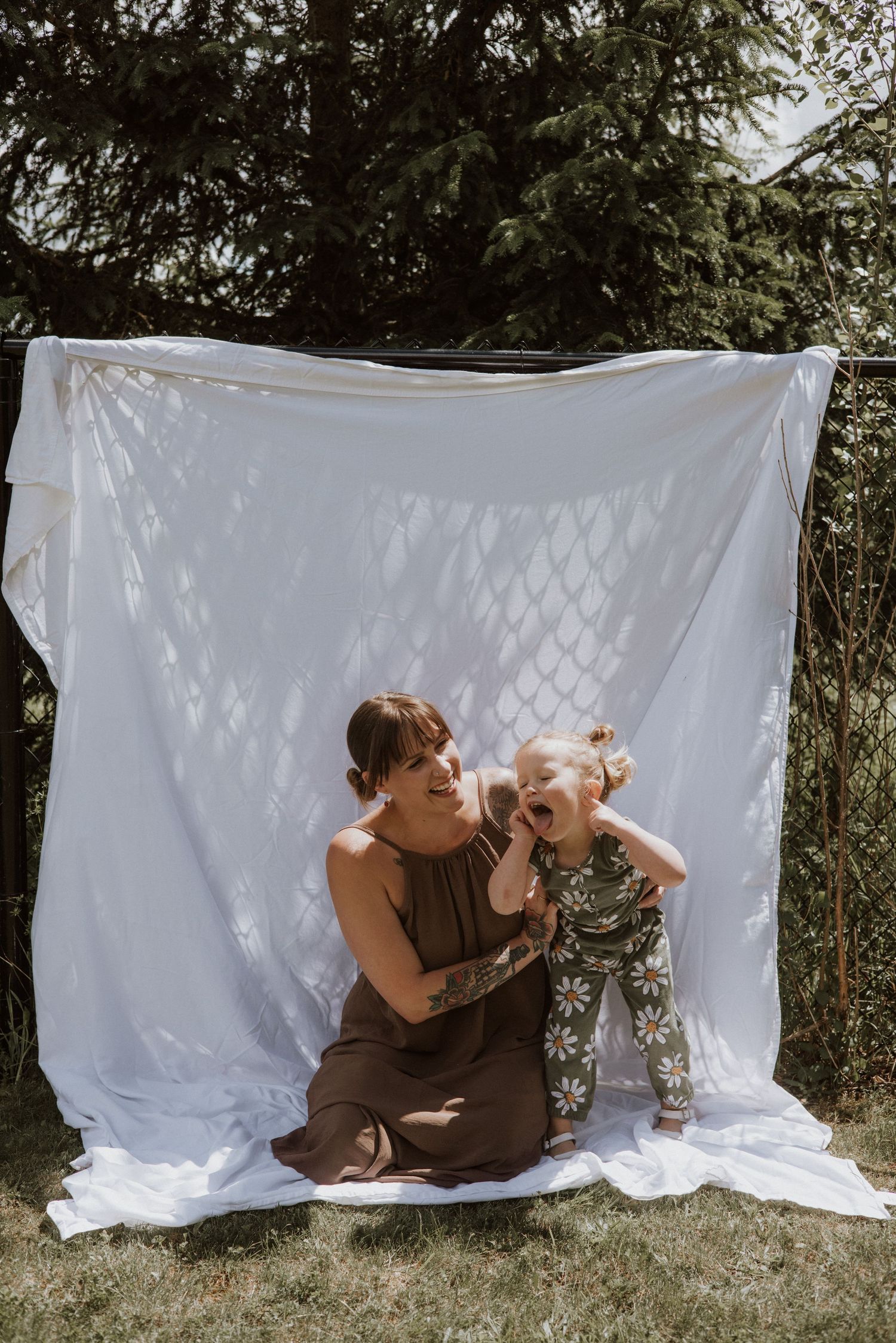 Mother in brown dress sitting on ground laughing with toddler daughter in daisy print outfit against white sheet backdrop.