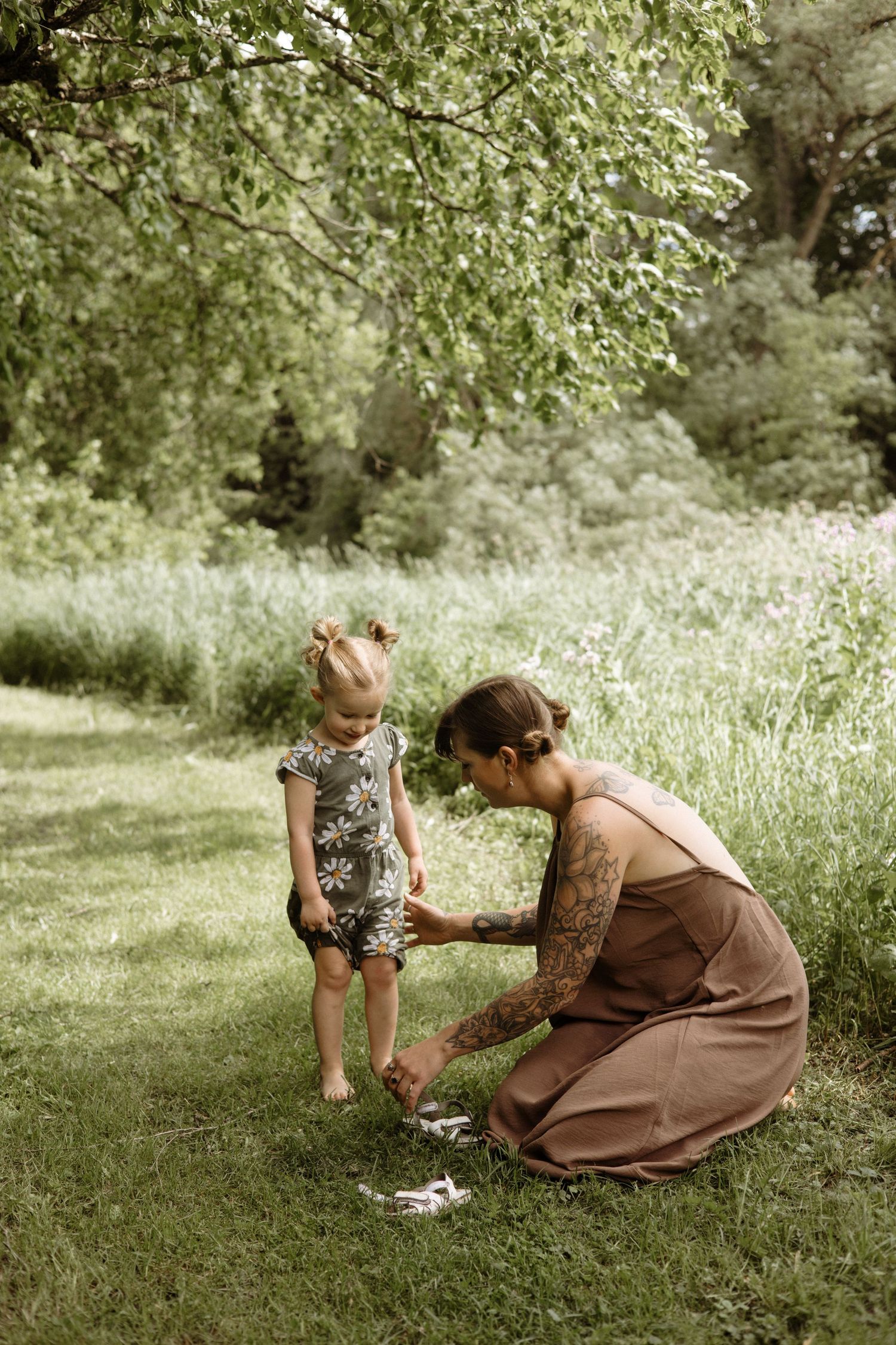 Tattooed mother in brown dress kneels on green grass helping young toddler daughter with something in her hands.