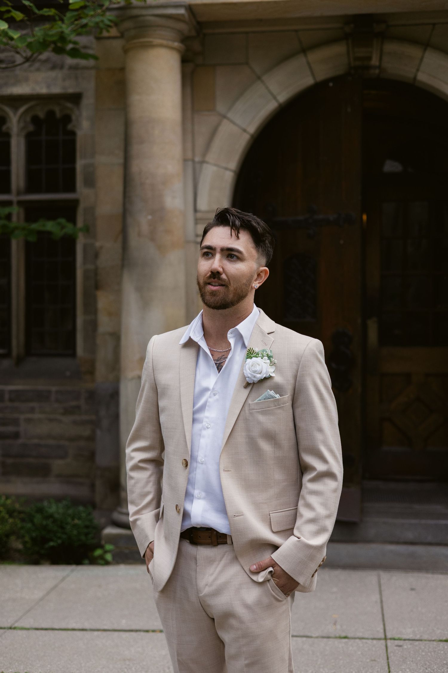 Groom in a beige linen suit with white boutonniere stands confidently before a stone archway entrance.