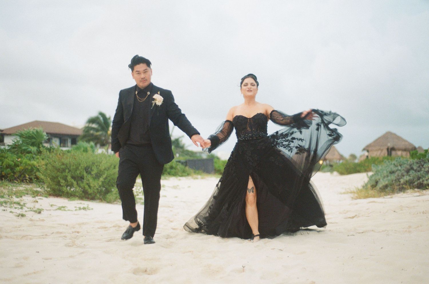 Stylish couple in all-black wedding attire walking hand in hand on a tropical beach.