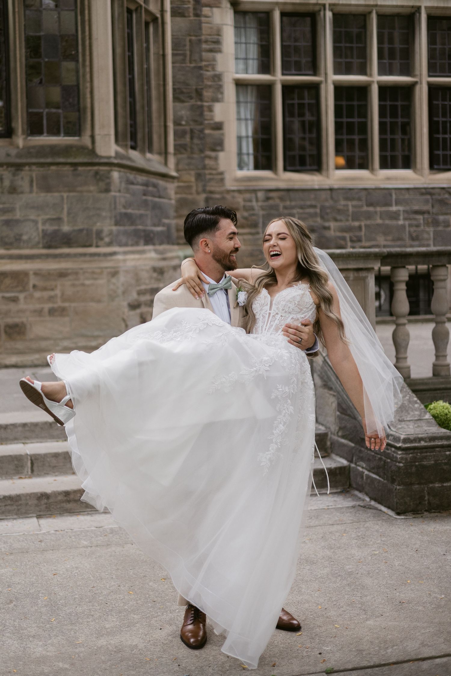 Groom lifts smiling bride in white lace gown outside a historic stone building on their wedding day.