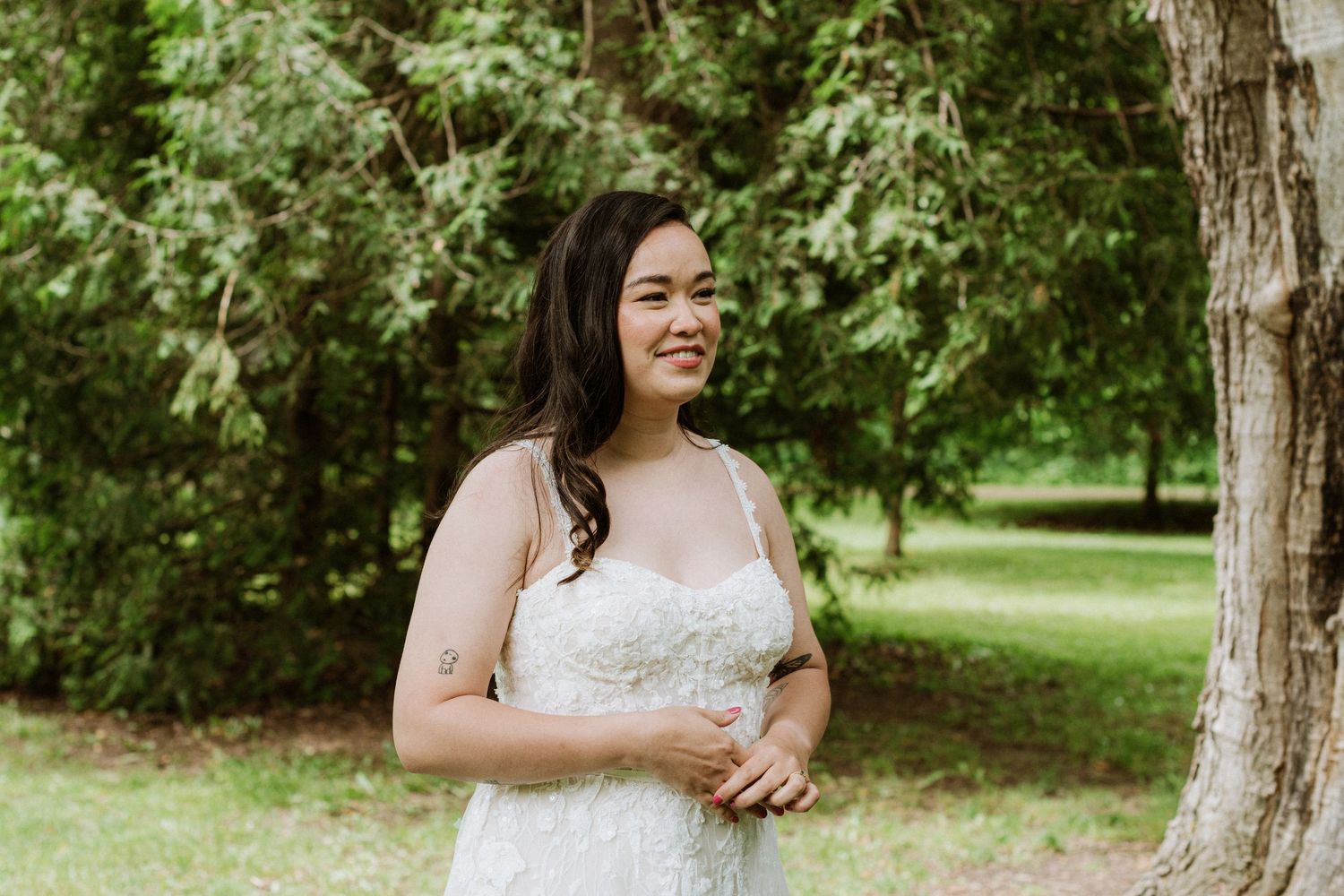 Smiling bride in strapless lace wedding gown stands outdoors near a tree in a green garden.