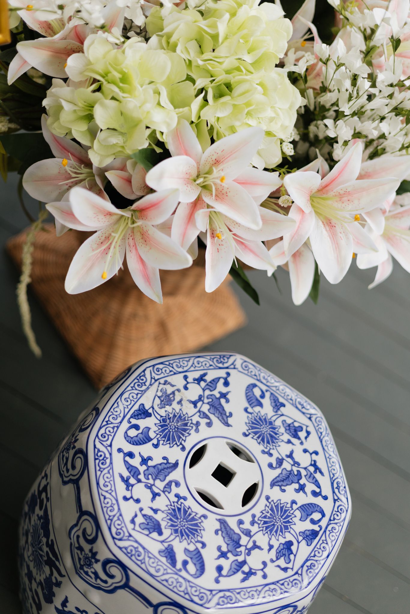 A blue and white chinoiserie garden stool beside a basket of pink lilies and hydrangeas.