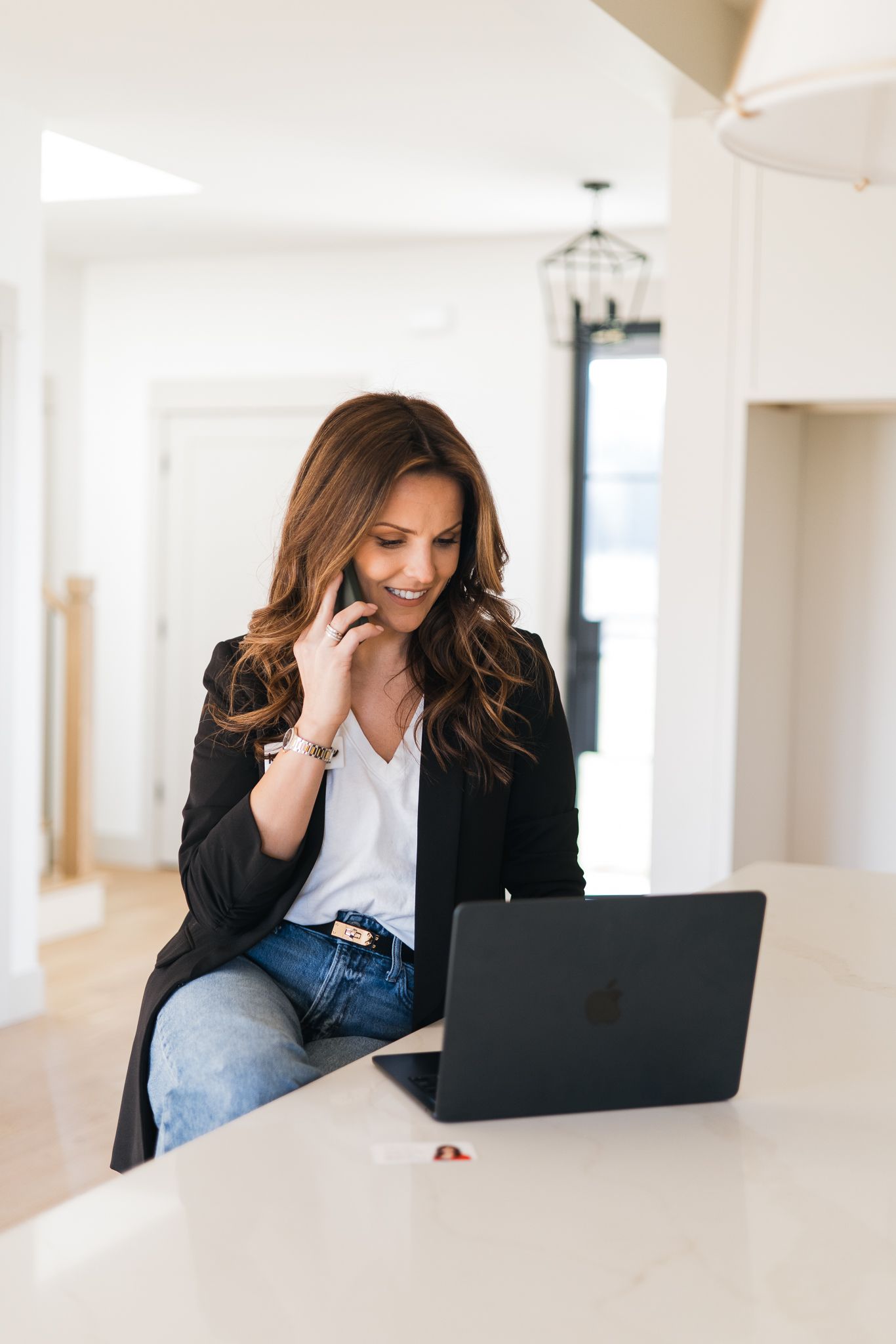 Brunette woman in black blazer talking on phone while working on laptop at a white kitchen counter.