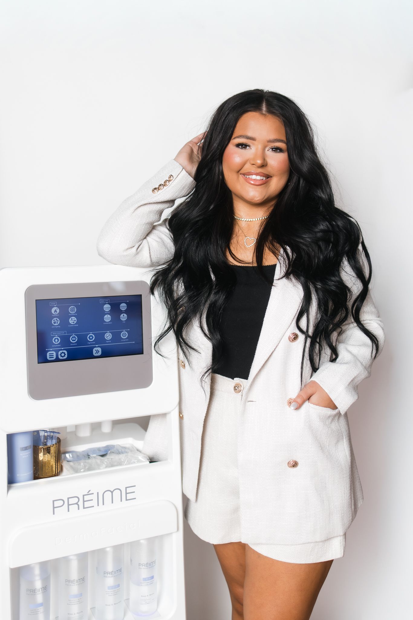 Smiling woman with long black hair in white blazer set posing beside Preime skincare device.
