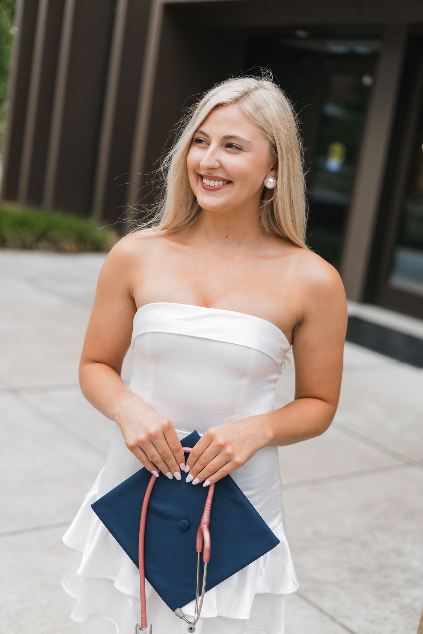 Smiling blonde graduate in white strapless dress holding navy blue graduation cap outdoors.