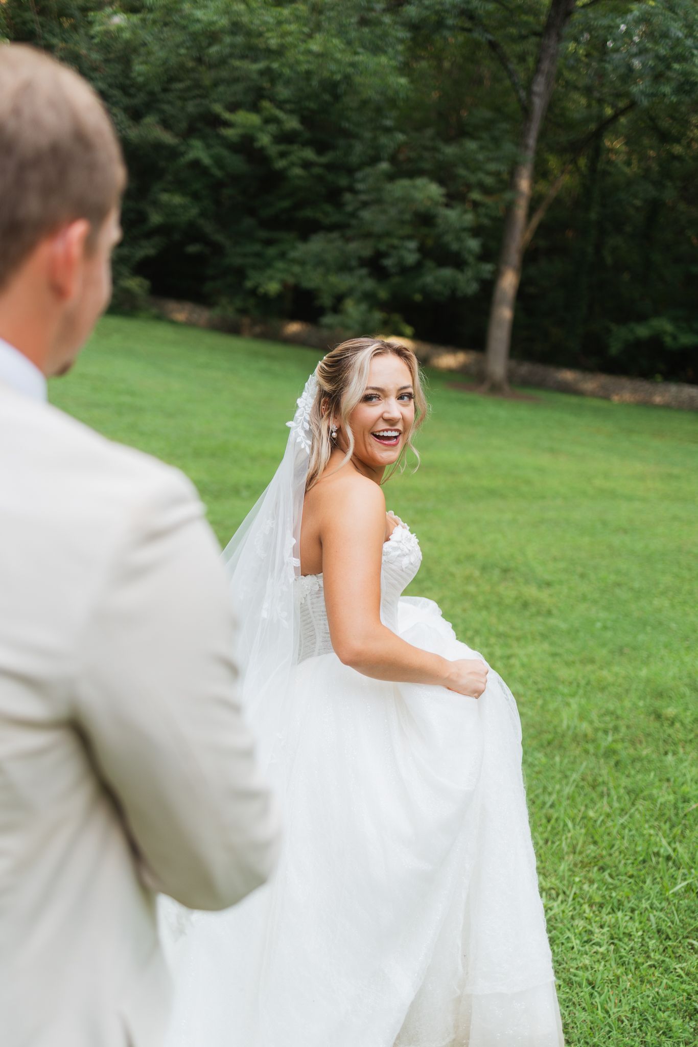 Smiling bride in white strapless gown and veil walks toward groom in tan suit on green lawn.