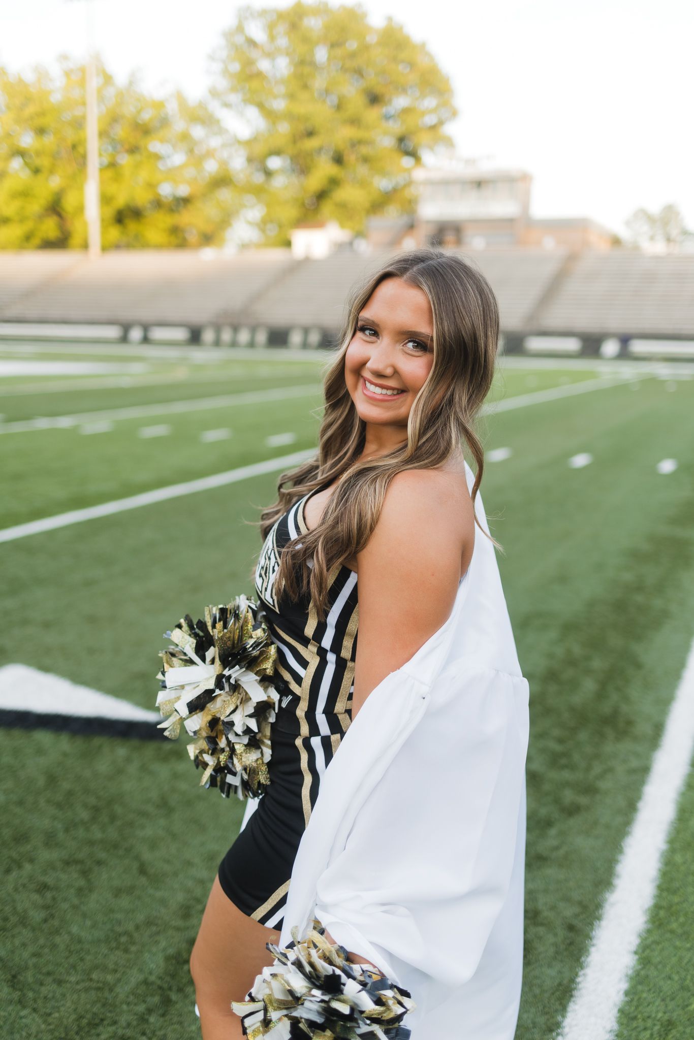 Cheerleader in black and gold uniform holds pom poms on football field while smiling over shoulder.