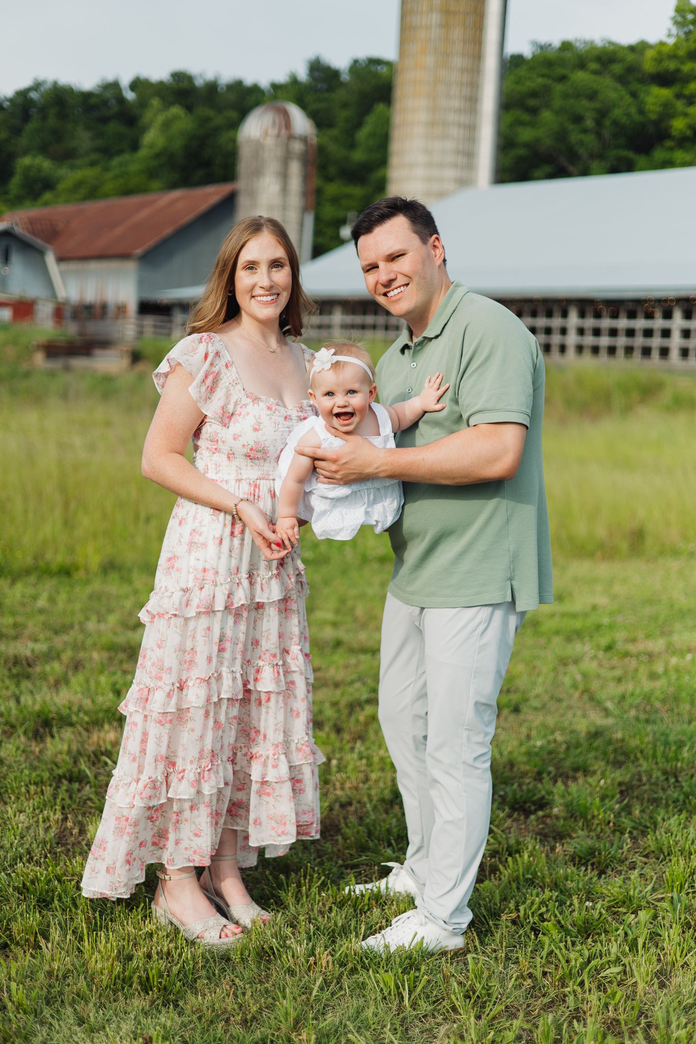 A smiling family of three poses in front of a farm barn, parents holding their laughing baby girl.