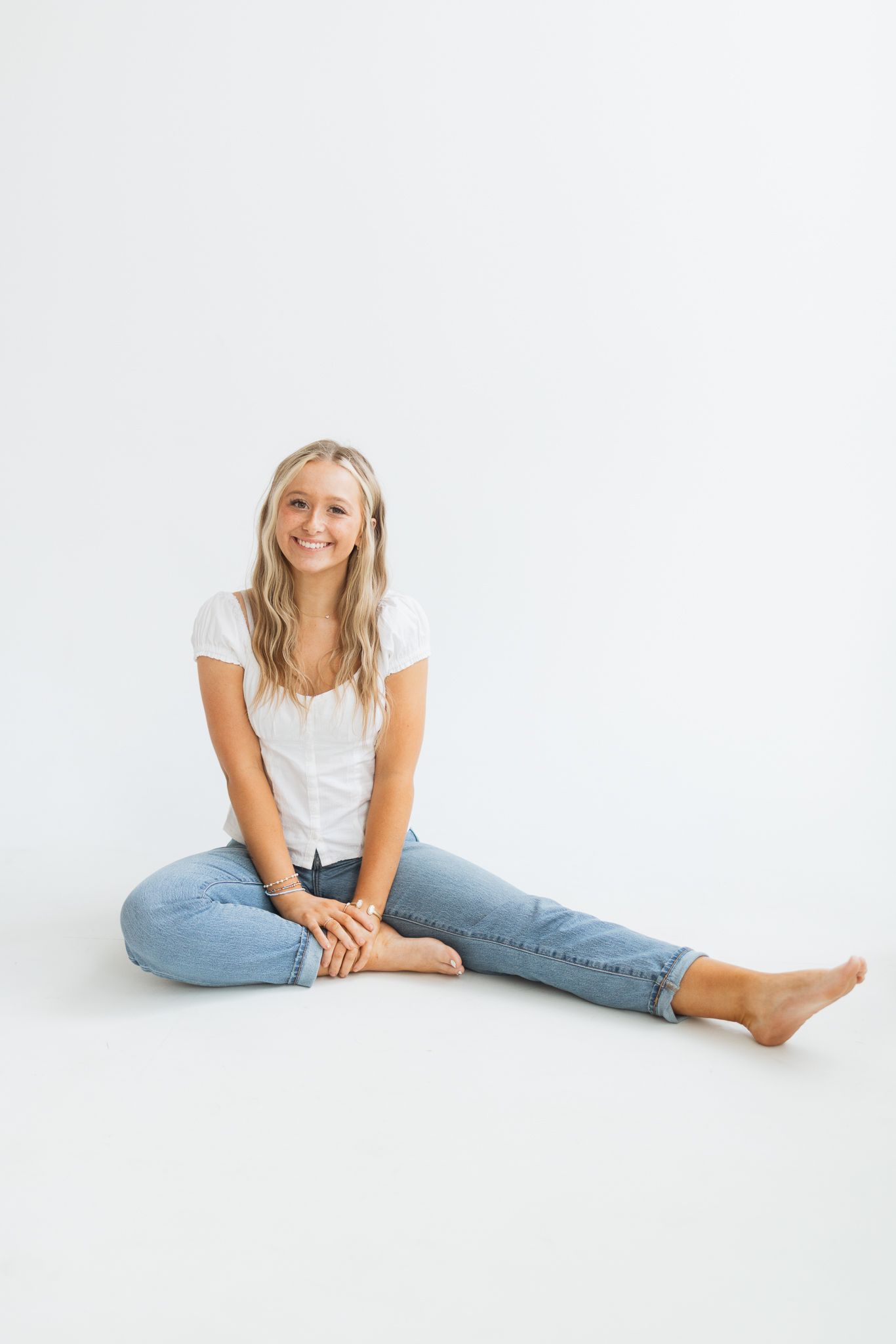 Smiling blonde woman sitting barefoot on white floor wearing white t-shirt and light blue jeans in studio setting.