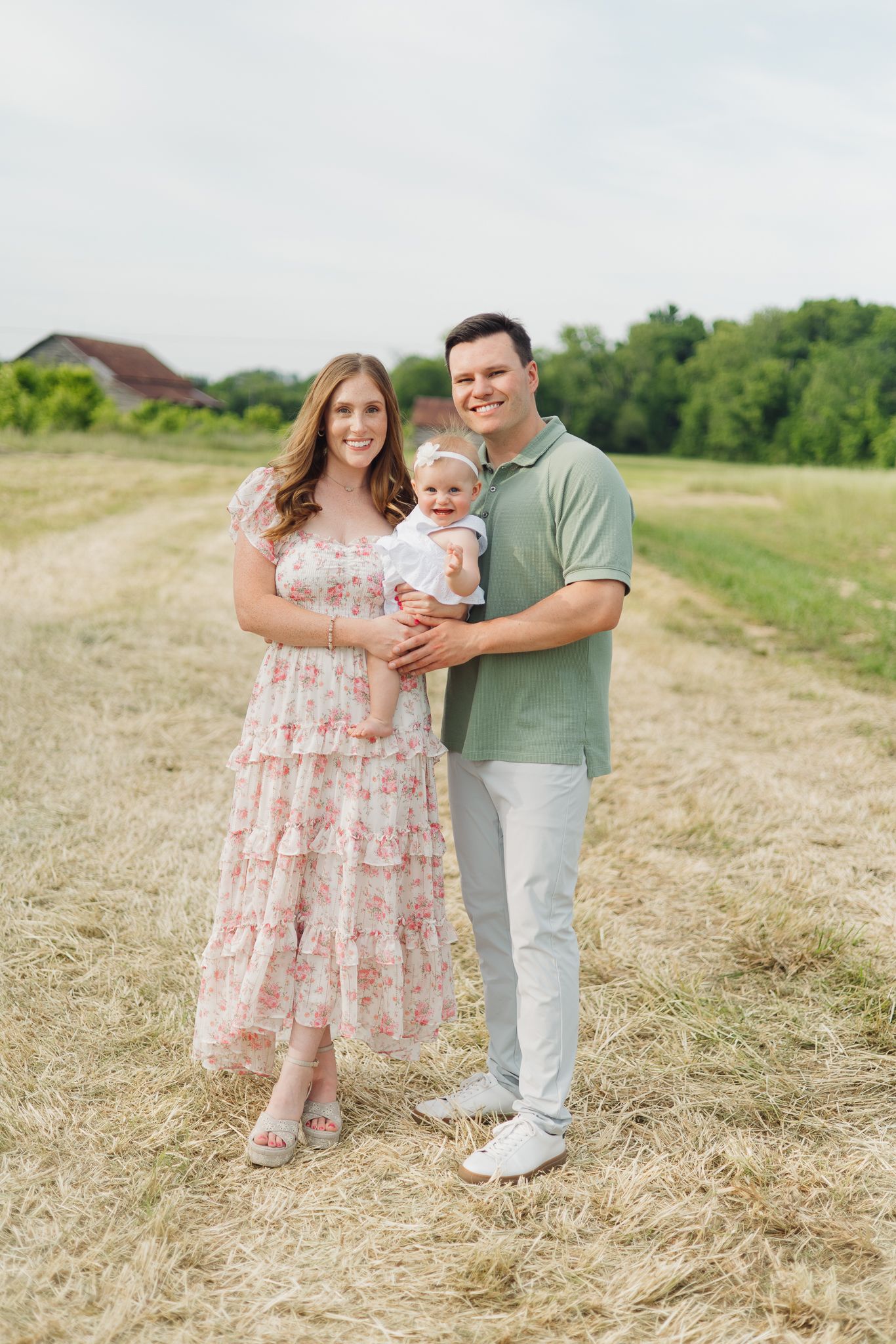 Happy young family with baby posing in a sunny field, mother in floral dress and father in green polo shirt.