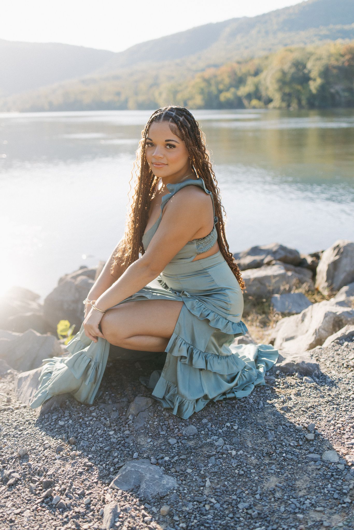 Young woman in sage green ruffled dress sitting on rocks by a serene lake with tree-covered hills in the background.