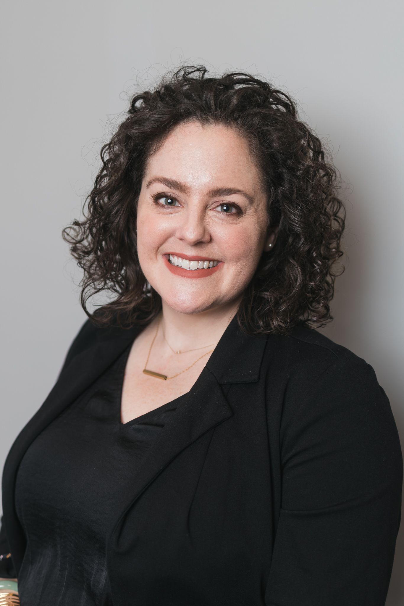 Professional woman with curly dark hair smiling, wearing a black blazer and gold necklace.