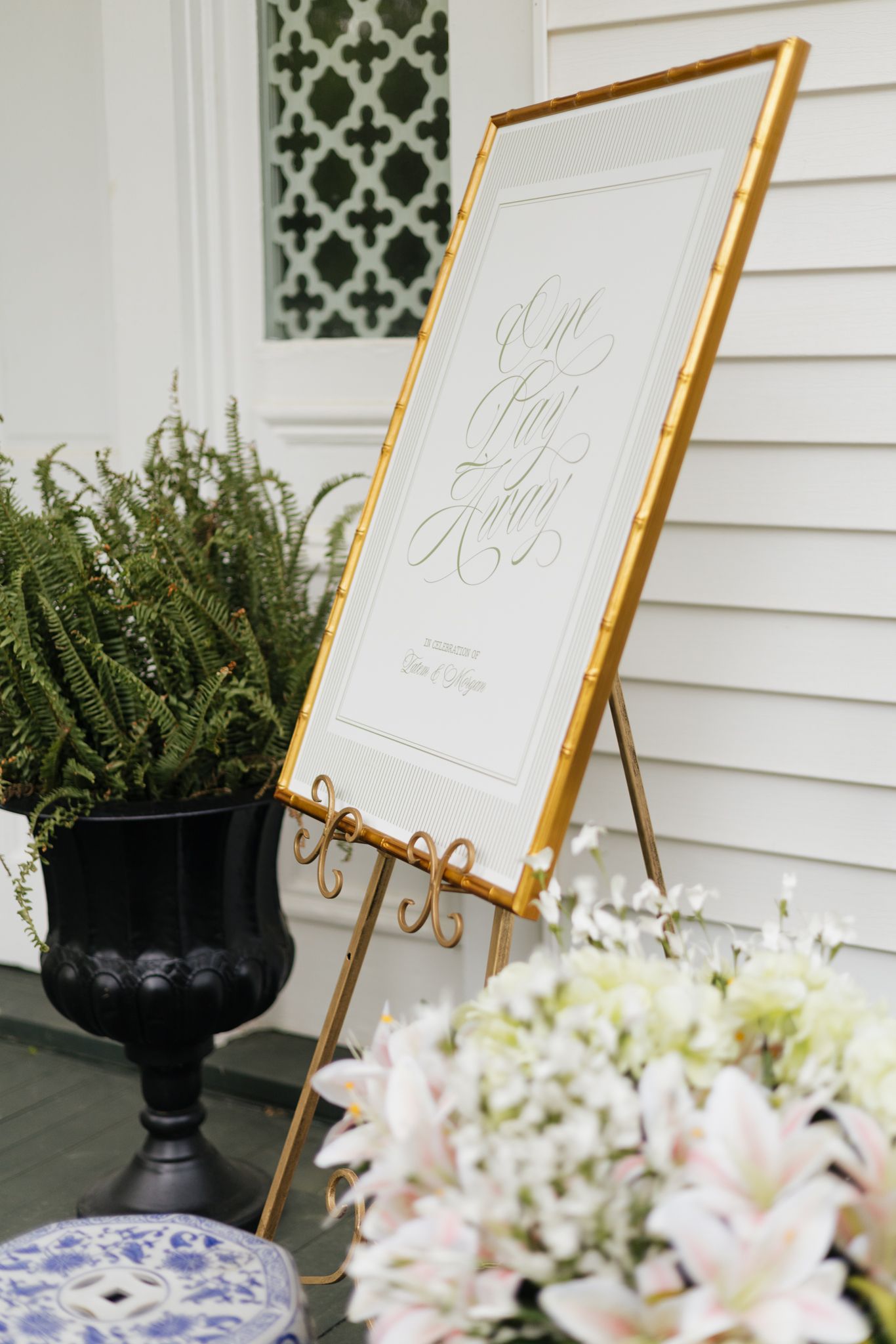 An elegant gold-framed wedding welcome sign on an easel surrounded by white lilies and ferns.