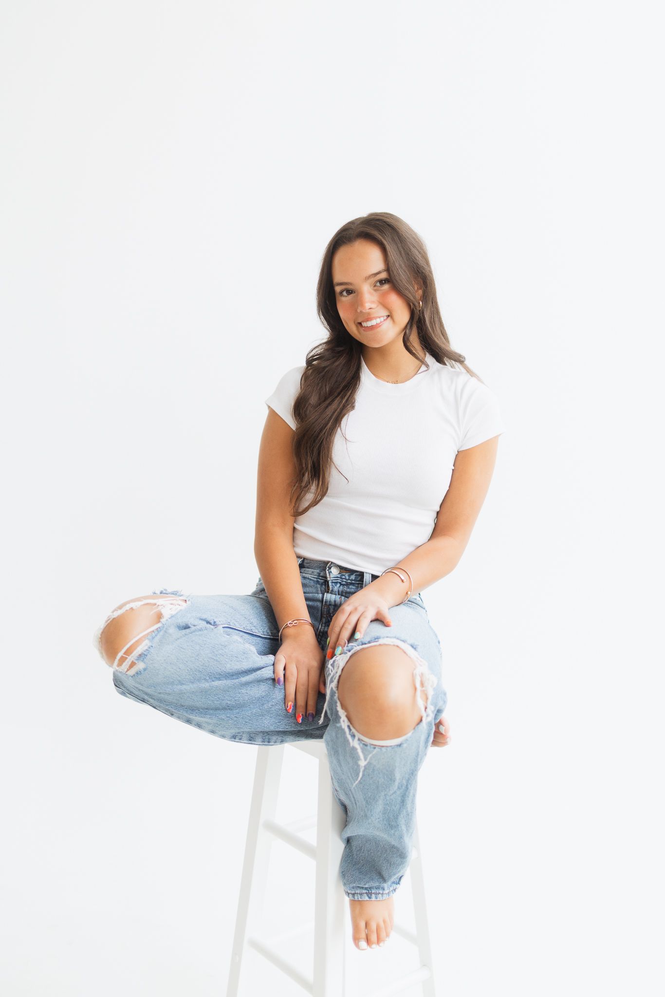 Young woman in white tee and ripped jeans sitting on a white stool, smiling against a white background.