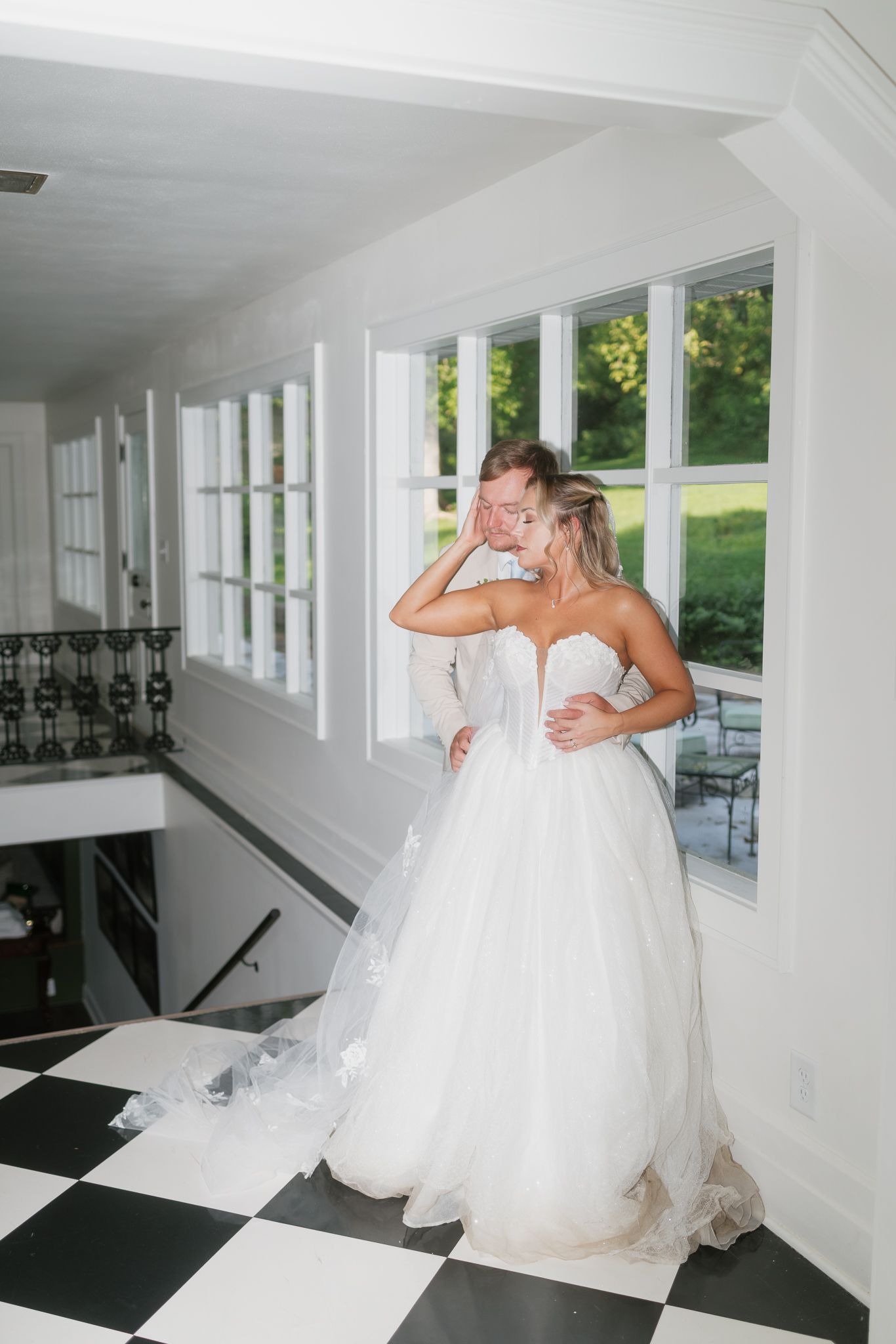 Two brides in white ball gowns share an emotional embrace by sunlit windows on a classic black and white checkered floor.