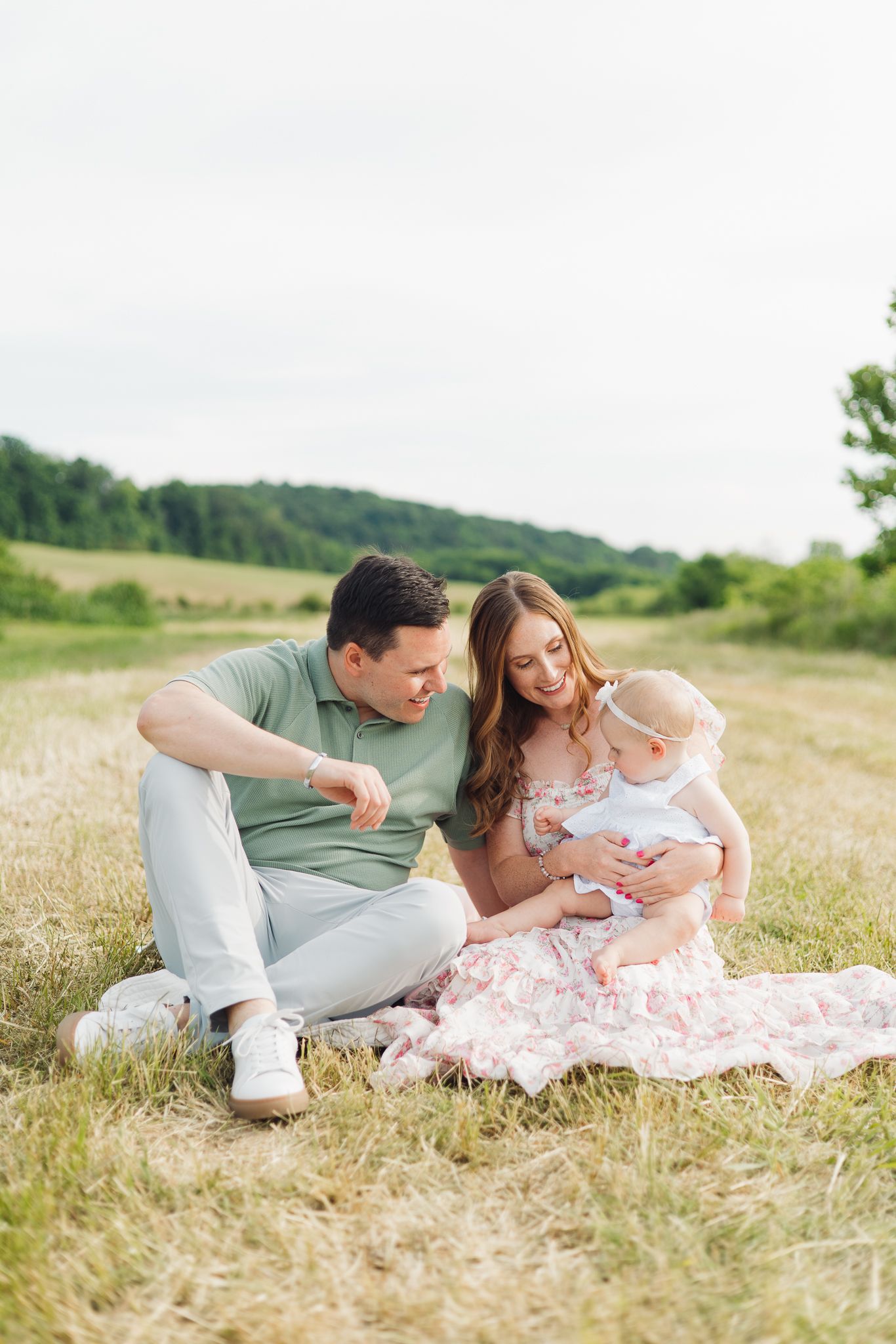 Young family sitting on a blanket in a field, parents smiling at their baby girl on a warm summer day.