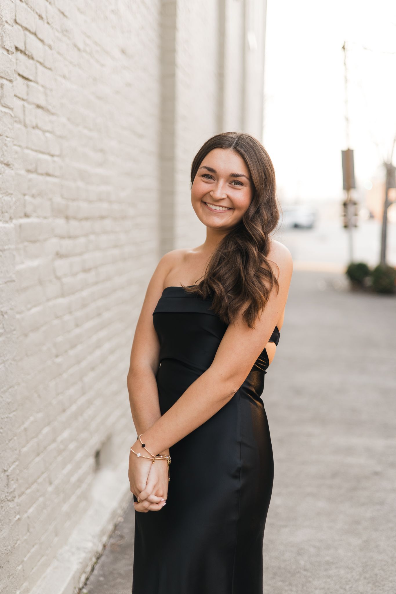 Smiling young woman in a black strapless satin gown posing in an urban alley beside a white brick wall.