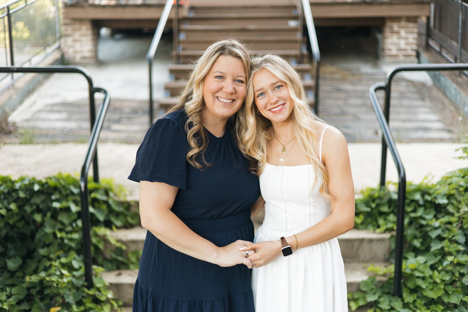 Two smiling women pose together outdoors on steps, one in navy dress, one in white sundress, surrounded by greenery.