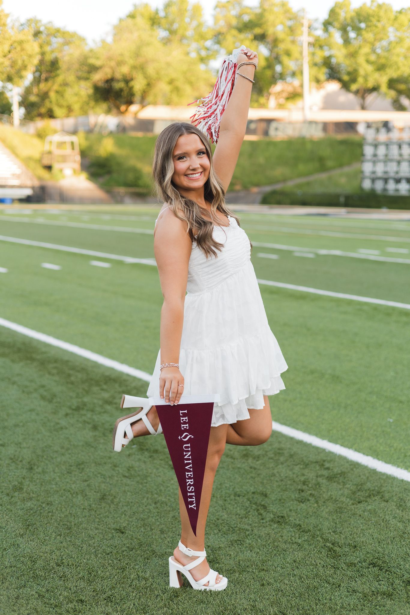 Smiling senior girl in white dress holds Lee University pennant and pom poms on football field.