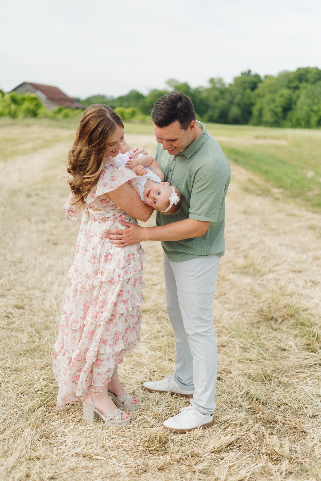A couple in a hay field gently passes their baby between them, sharing a tender family moment.
