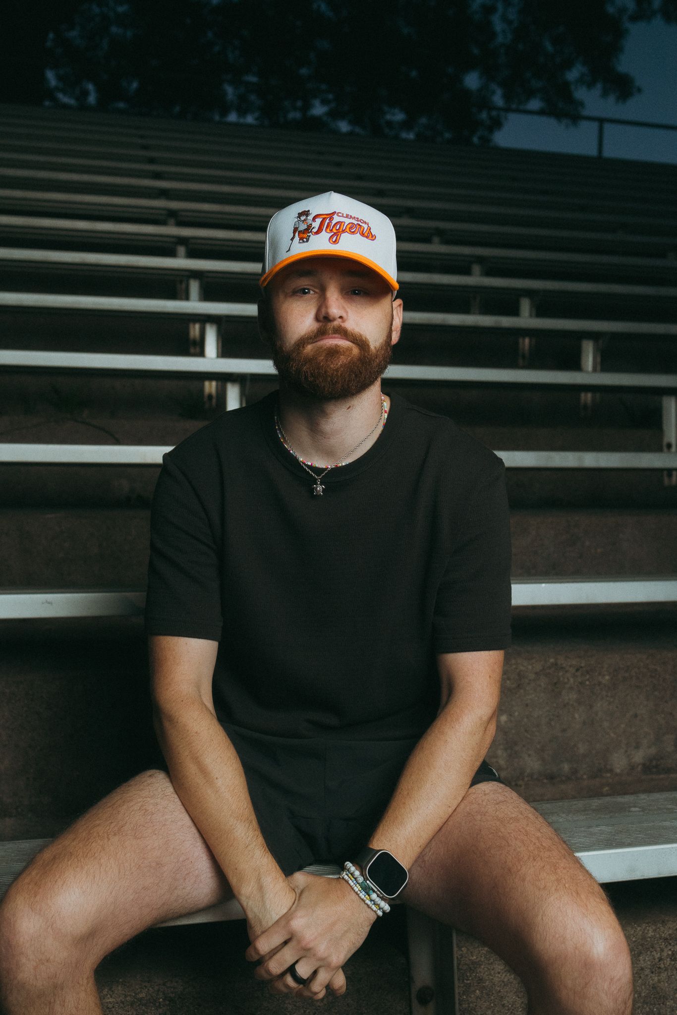 Bearded man wearing a Detroit Tigers snapback cap sitting on stadium bleachers.