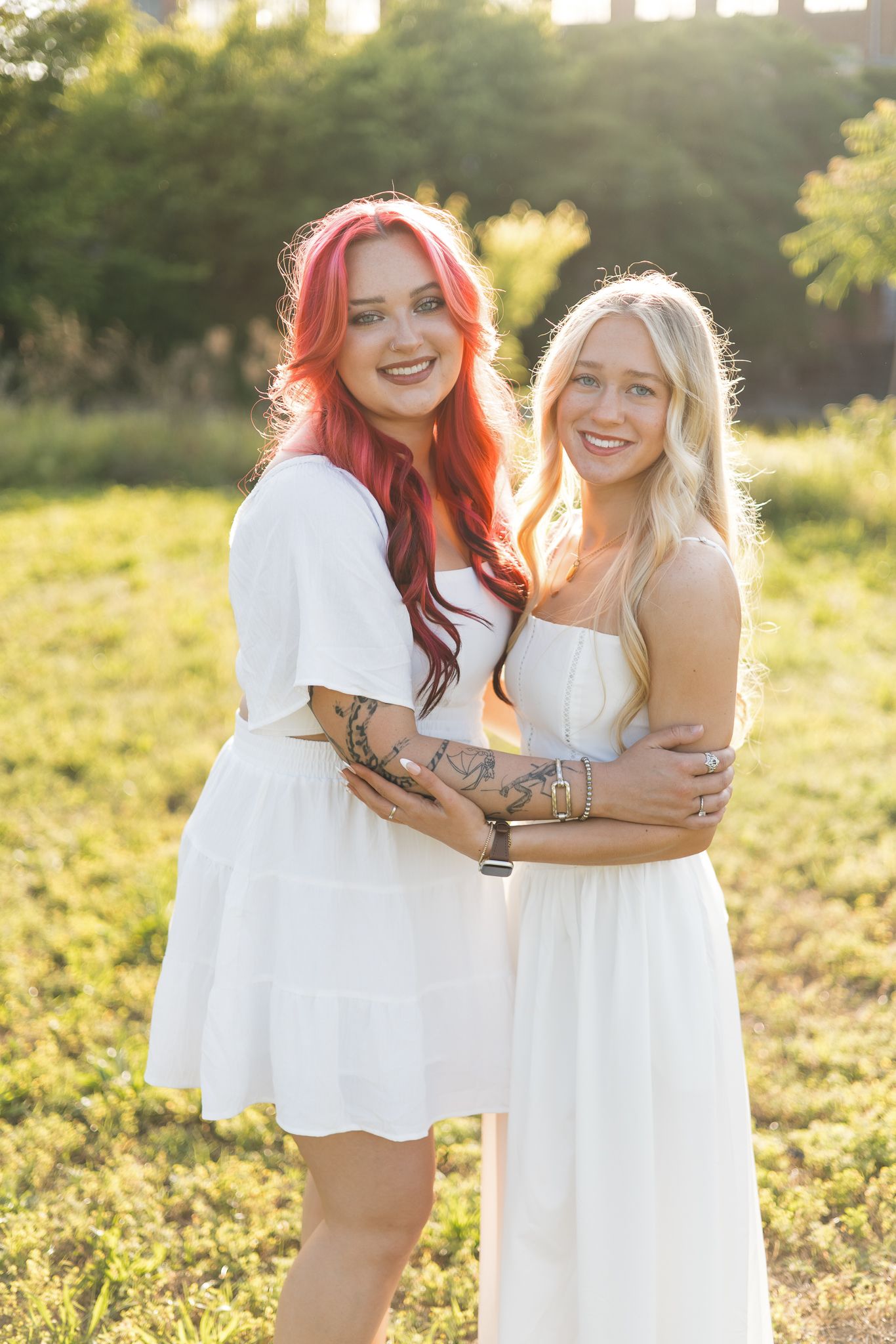 Two smiling women in white dresses embracing outdoors, one with red hair and tattoos, in golden sunlight.