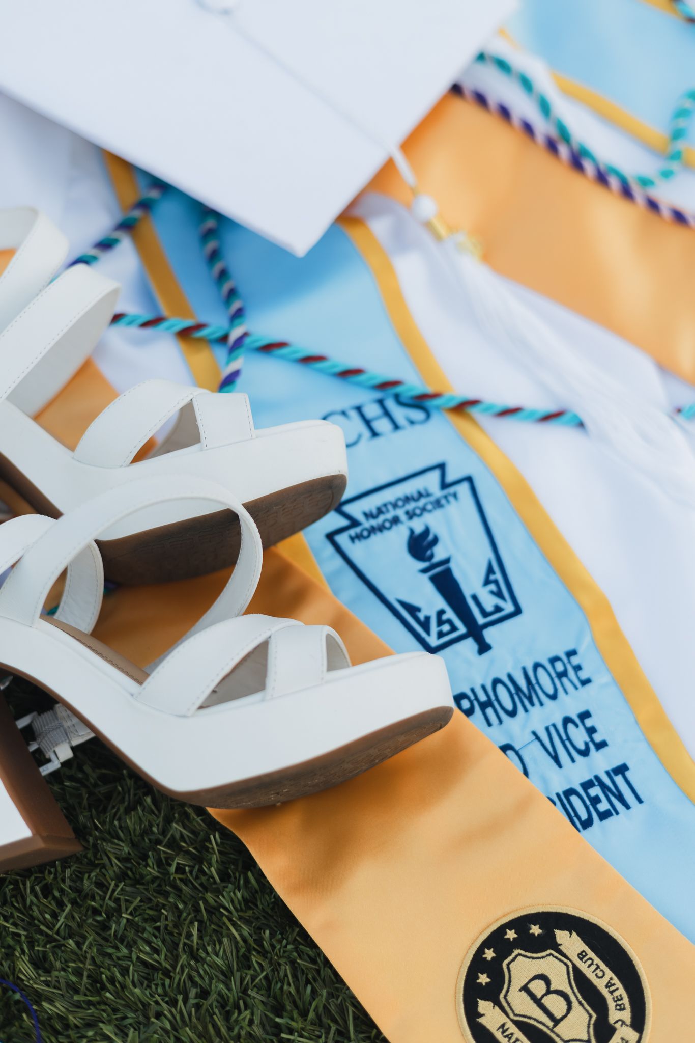 White platform heels rest on National Honor Society graduation stole and academic regalia on grass.