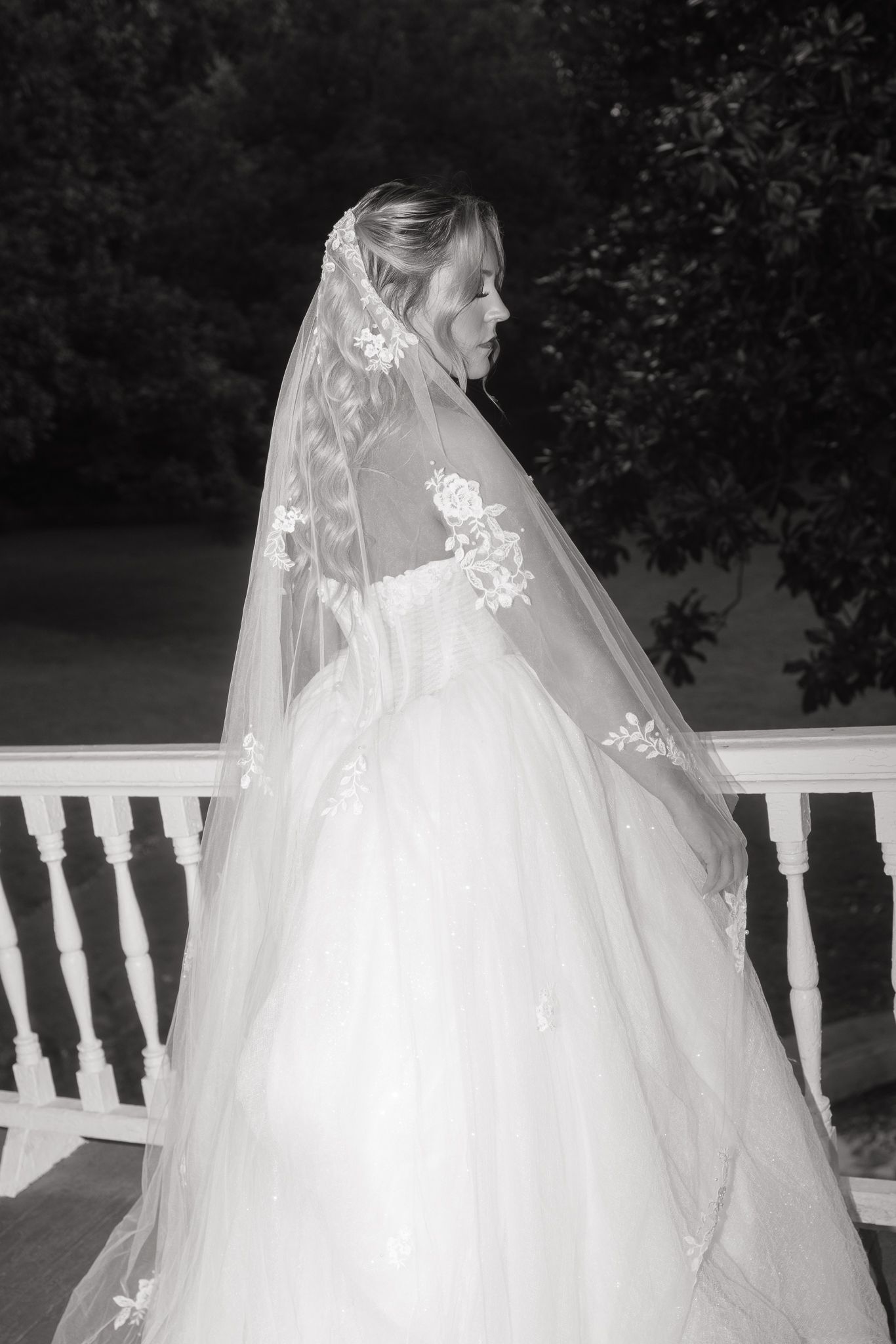 Black and white photo of a bride in a ballgown with lace veil on a porch.
